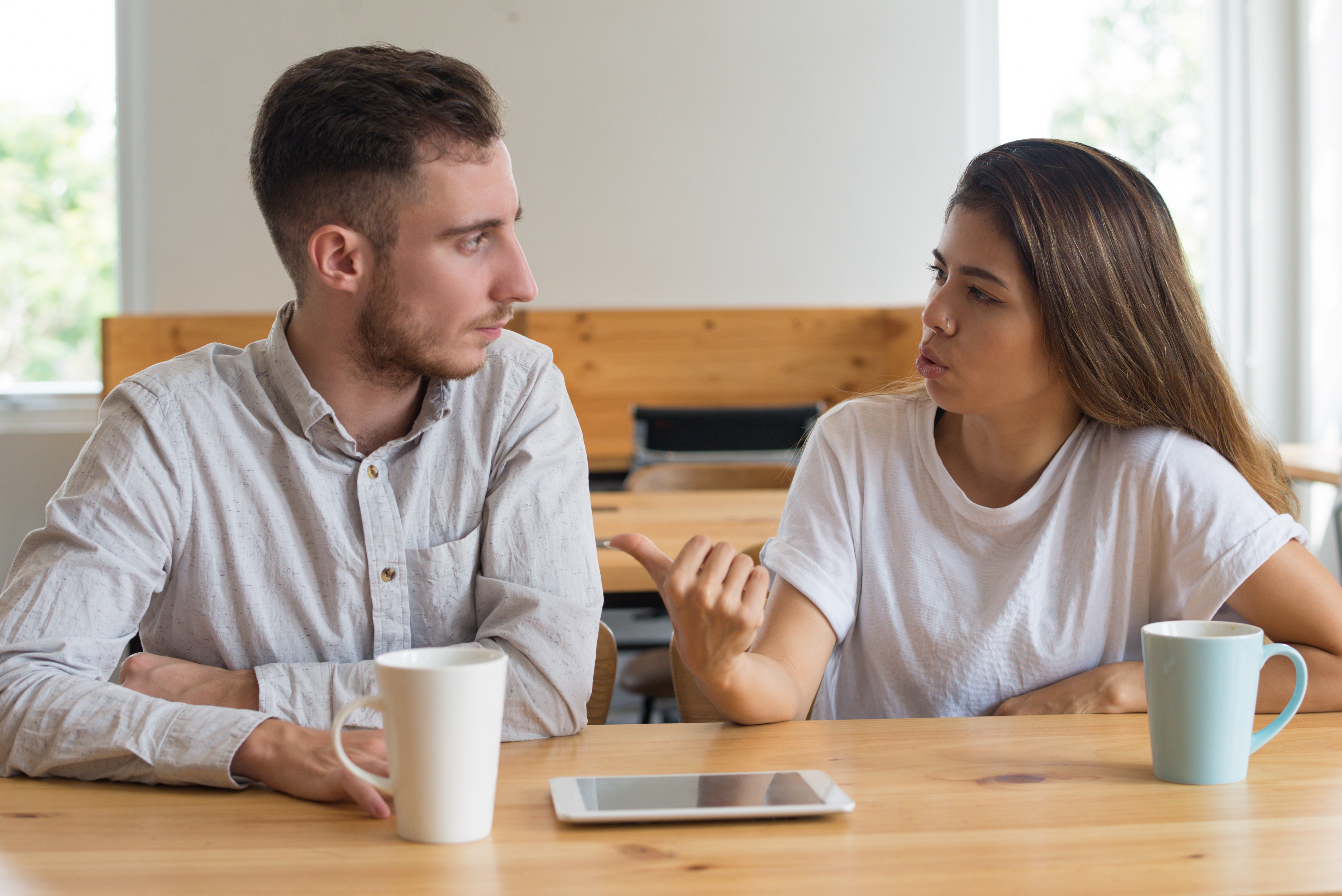 Homem e mulher jovens sentados &agrave; mesa de madeira, conversando. Ele, de camisa clara, olha para ela, que gesticula com a m&atilde;o direita, vestindo camiseta branca. H&aacute; canecas e um tablet sobre a mesa