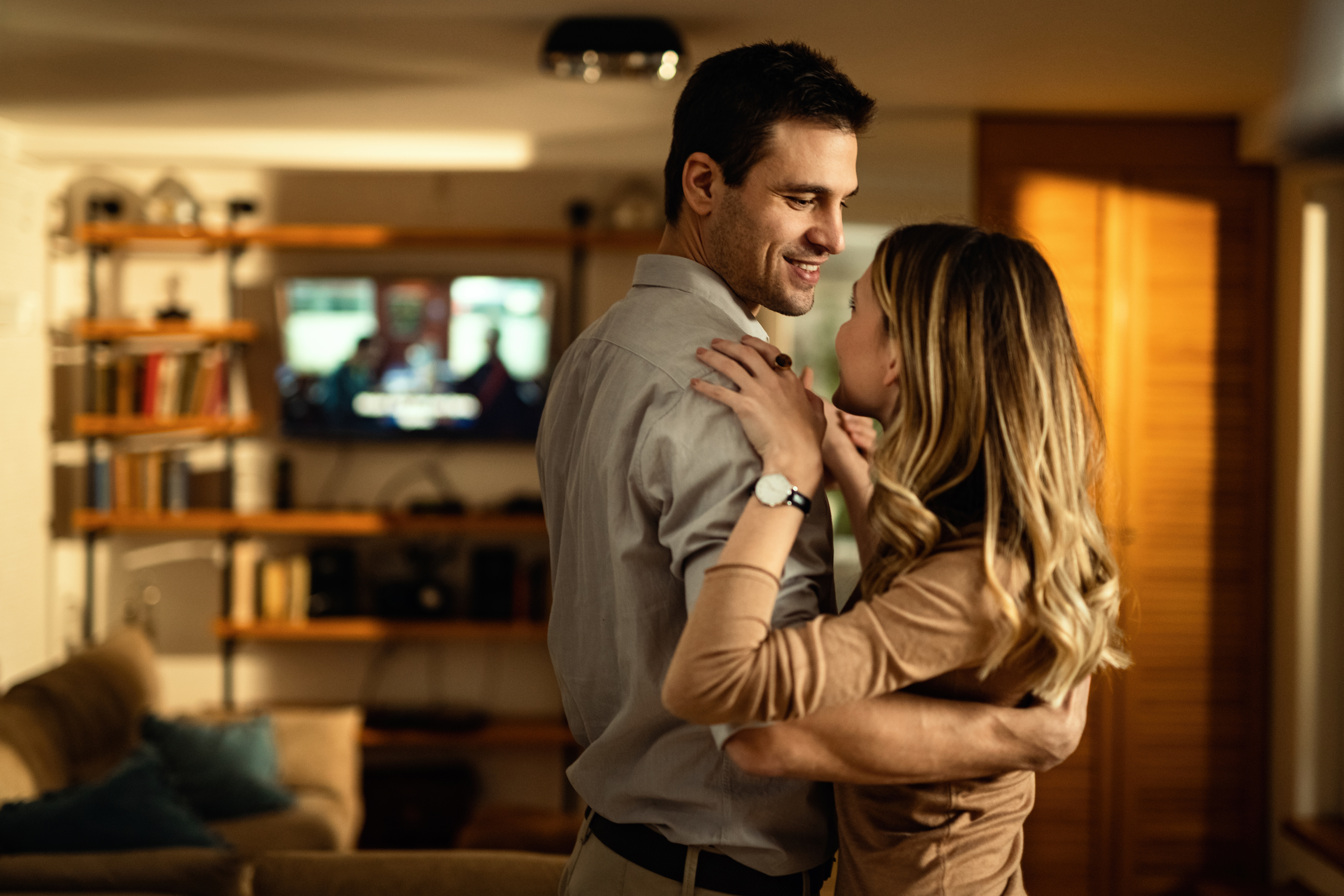 Casal jovem e sorridente se abraça em uma sala de estar aconchegante, com a mulher de cabelos longos loiros olhando para o homem, que veste camisa cinza. Ao fundo, uma estante com livros e uma televisão ligada.