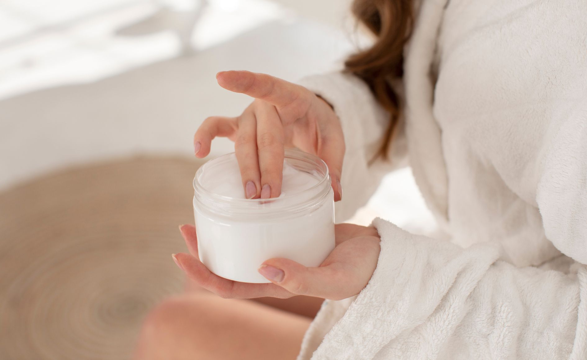 Mãos de uma mulher com roupão branco pegando creme hidratante de um pote transparente, em um ambiente claro