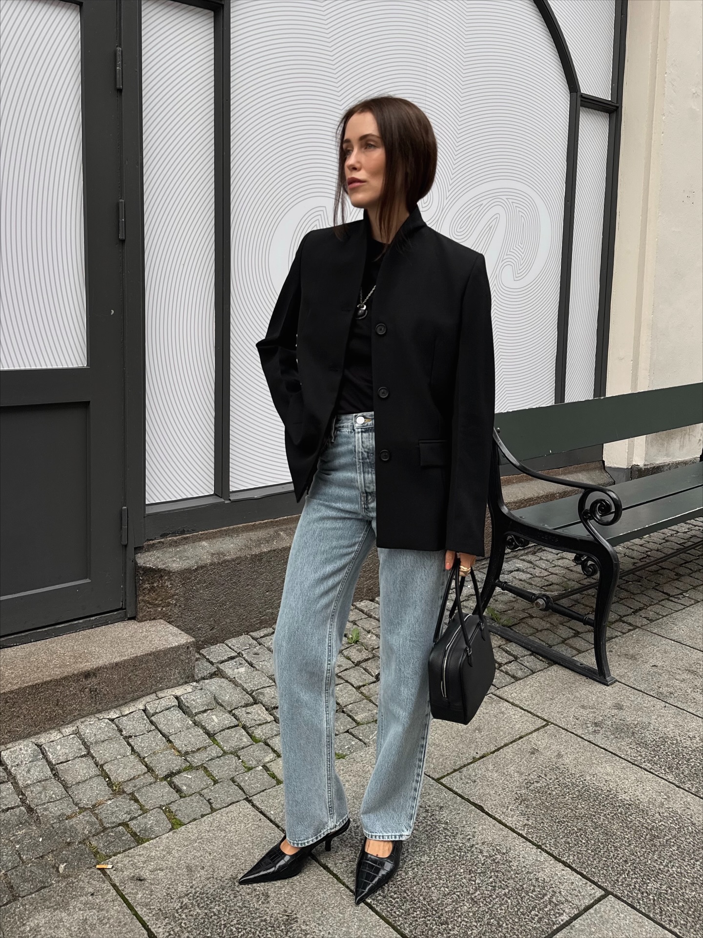 Mulher de cabelo castanho, blazer preto, camiseta preta, cal&ccedil;a jeans clara e sapatos pretos de bico fino, segurando uma bolsa preta, em p&eacute; na cal&ccedil;ada de paralelep&iacute;pedos, olhando para a esquerda. Ao fundo, uma porta escura e uma parede branca com padr&otilde;es circulares, al&eacute;m de um banco de pra&ccedil;a verde escuro