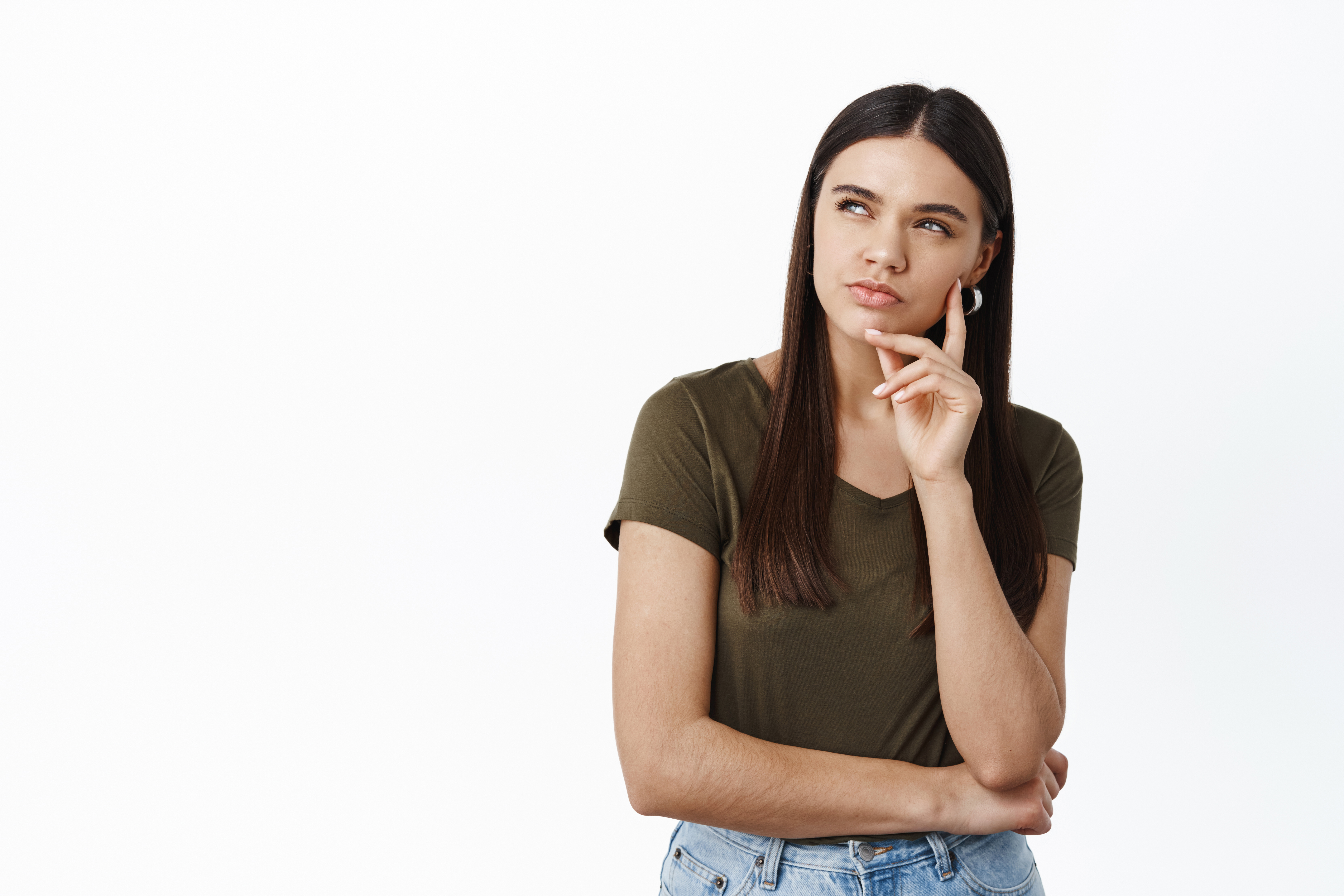 Mulher jovem com cabelo castanho escuro, camiseta verde-oliva e jeans, pensativa, com a m&atilde;o no queixo e olhando para cima, &agrave; esquerda, sobre fundo branco