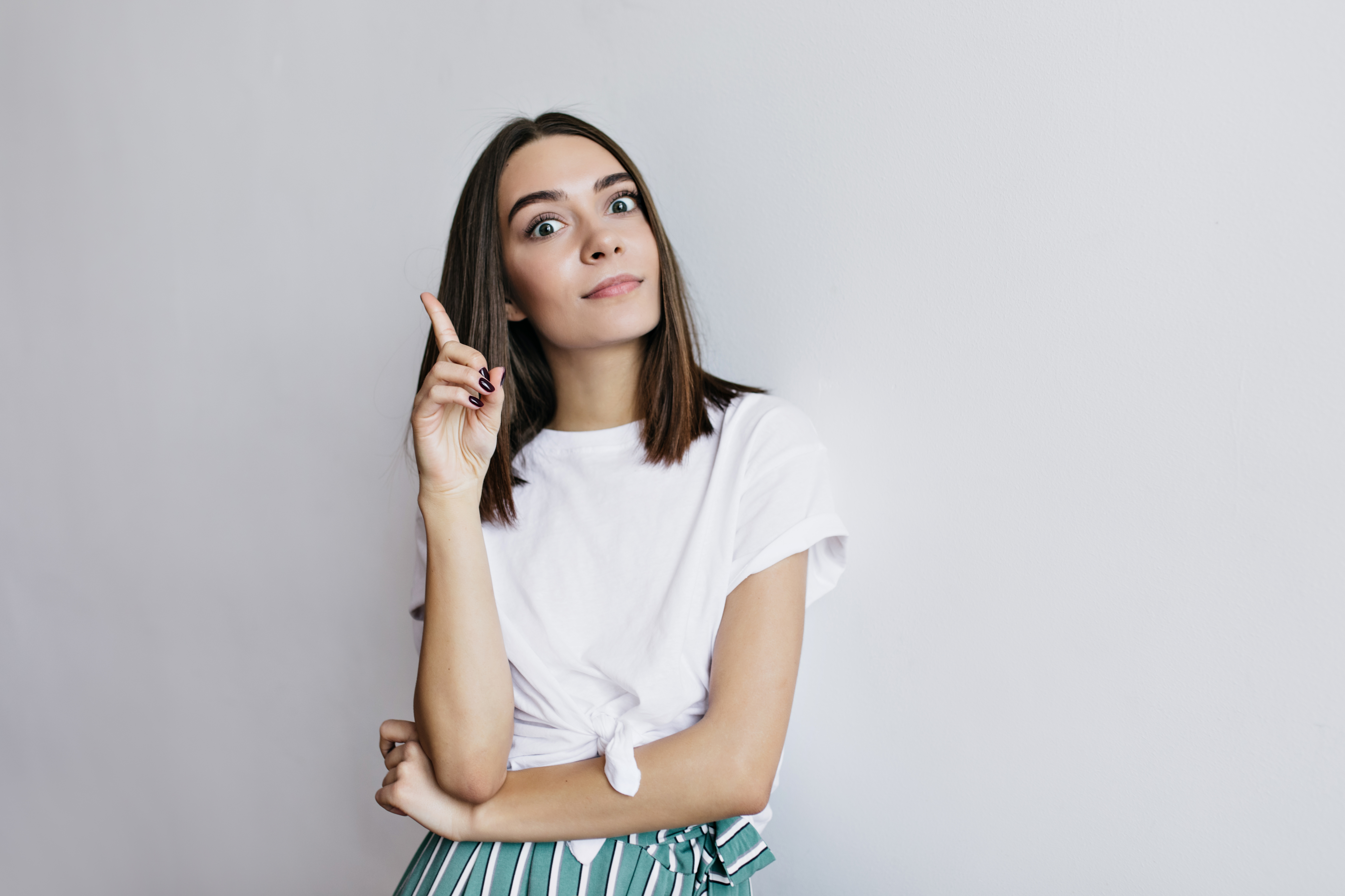 Mulher jovem, de cabelo castanho e liso, com olhos arregalados e boca levemente aberta, apontando o dedo indicador para cima. Veste camiseta branca com n&oacute; na barra e saia listrada verde e branca, com o bra&ccedil;o esquerdo cruzado sobre o corpo. Fundo cinza claro
