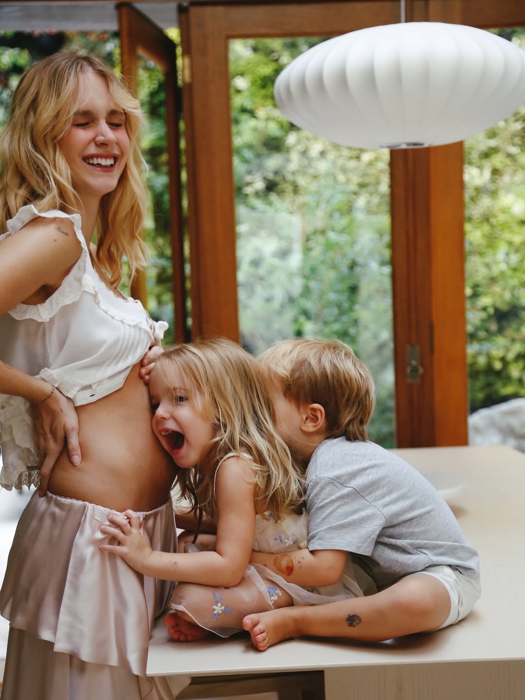 Mulher gr&aacute;vida sorrindo, com dois filhos pequenos, uma menina e um menino, abra&ccedil;ando sua barriga em um ambiente iluminado com janelas e plantas ao fundo