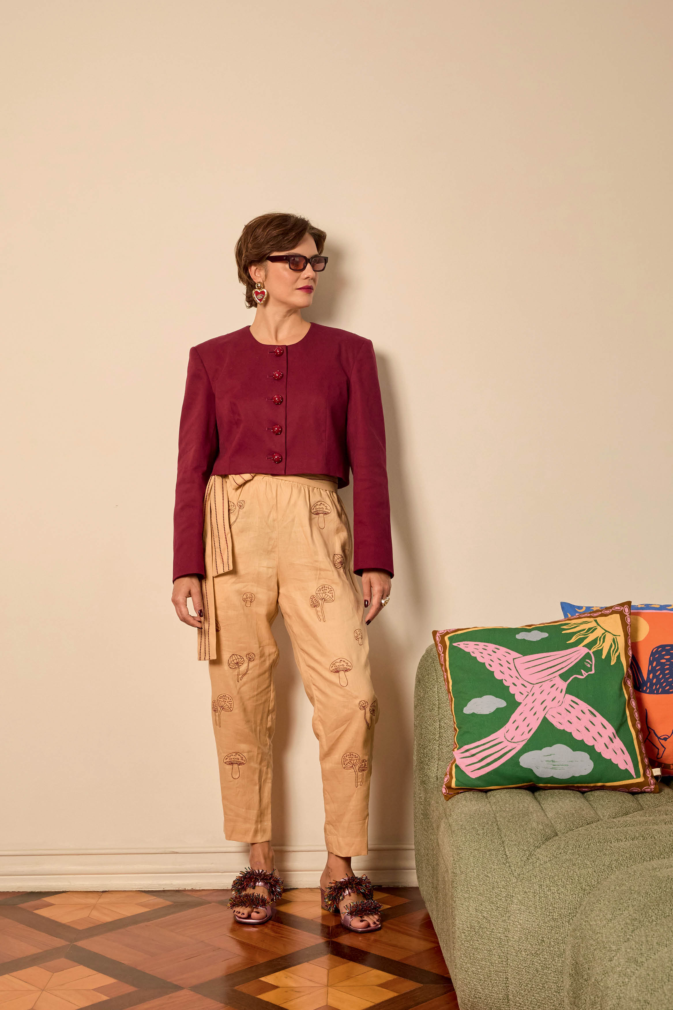 Mulher de pele clara, cabelo curto castanho, &oacute;culos escuros e brincos de cora&ccedil;&atilde;o vermelho, vestindo blazer vinho e cal&ccedil;a bege com estampa de cogumelos, posa em p&eacute; ao lado de um sof&aacute; verde com almofadas coloridas