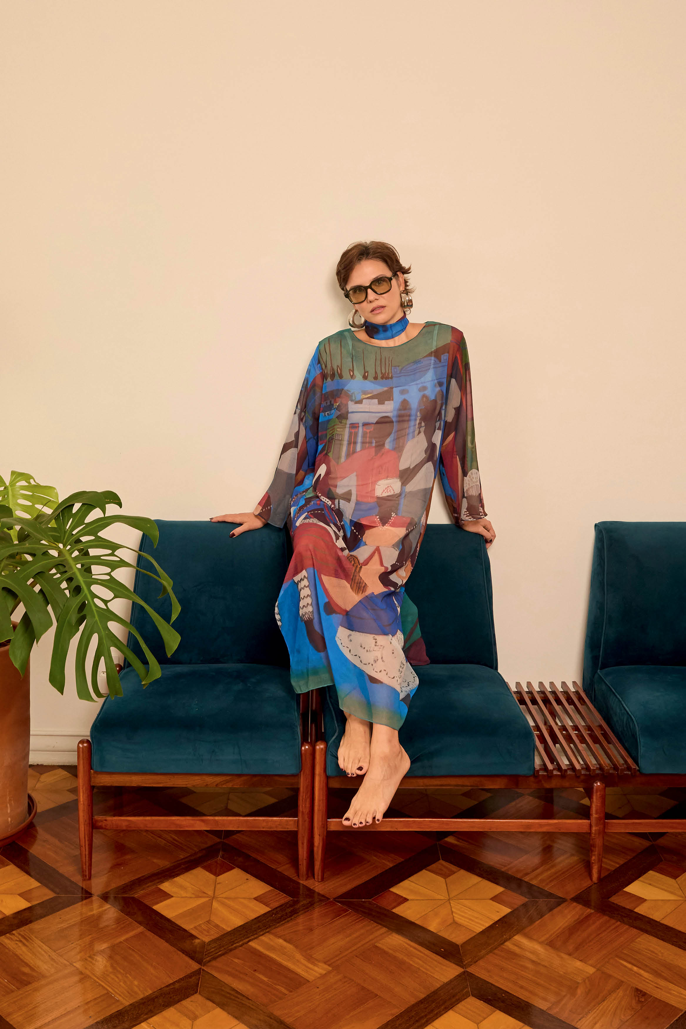 Mulher de &oacute;culos escuros e cabelo curto, vestindo um longo vestido estampado com figuras abstratas coloridas, sentada descal&ccedil;a no encosto de um sof&aacute; azul-petr&oacute;leo, com uma planta verde &agrave; esquerda e piso de madeira com padr&otilde;es geom&eacute;tricos