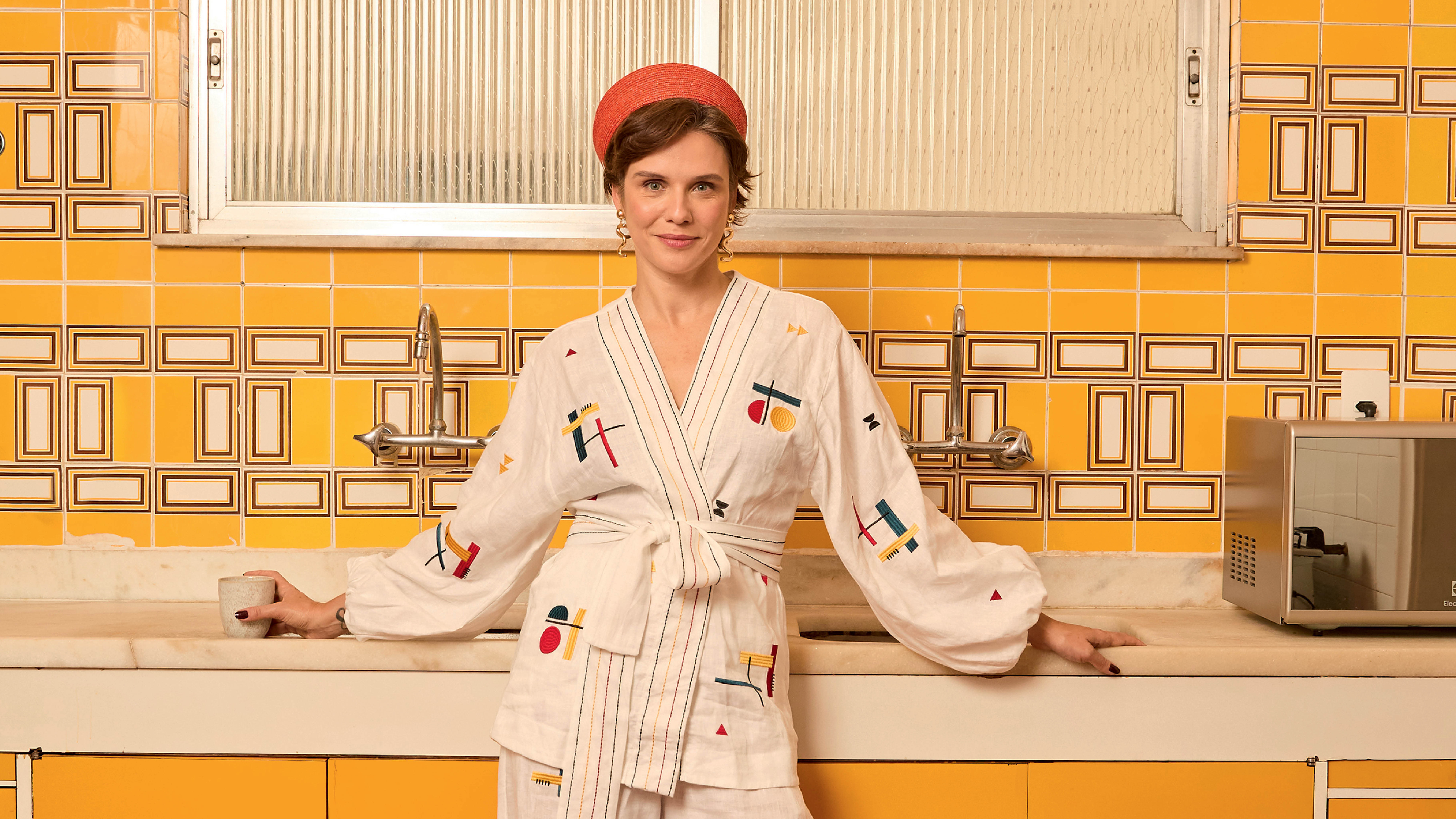 Mulher sorridente com chapéu laranja e quimono branco estampado, apoiada em balcão de cozinha com azulejos amarelos e pia. Ela segura uma caneca na mão esquerda e um micro-ondas está à direita