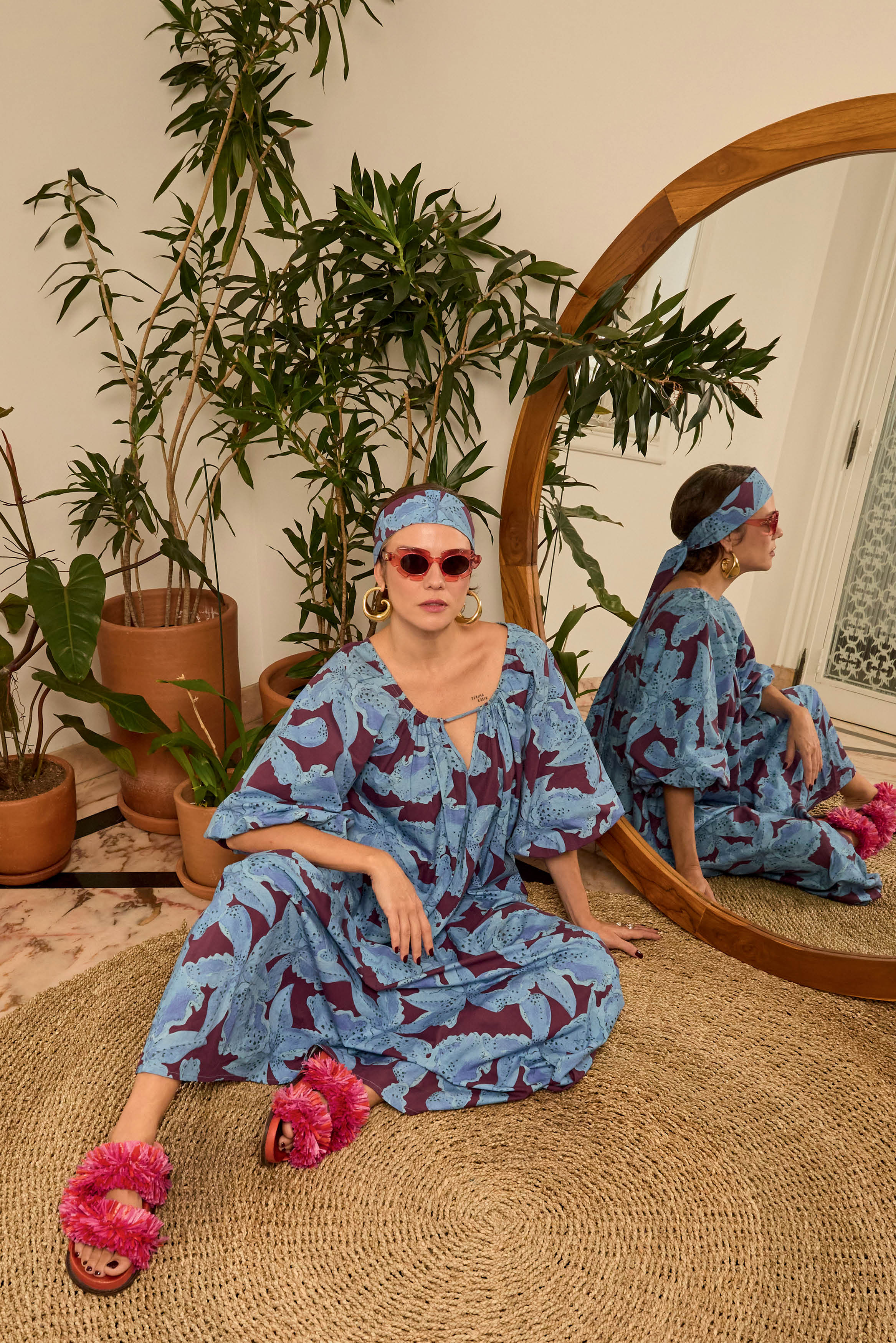 Mulher sentada no ch&atilde;o sobre tapete de sisal, usando vestido azul e vinho estampado, faixa de cabelo combinando, &oacute;culos de sol rosa e sand&aacute;lias pink de pompons. Ao fundo, plantas em vasos de barro e um espelho redondo de madeira refletindo seu perfil.