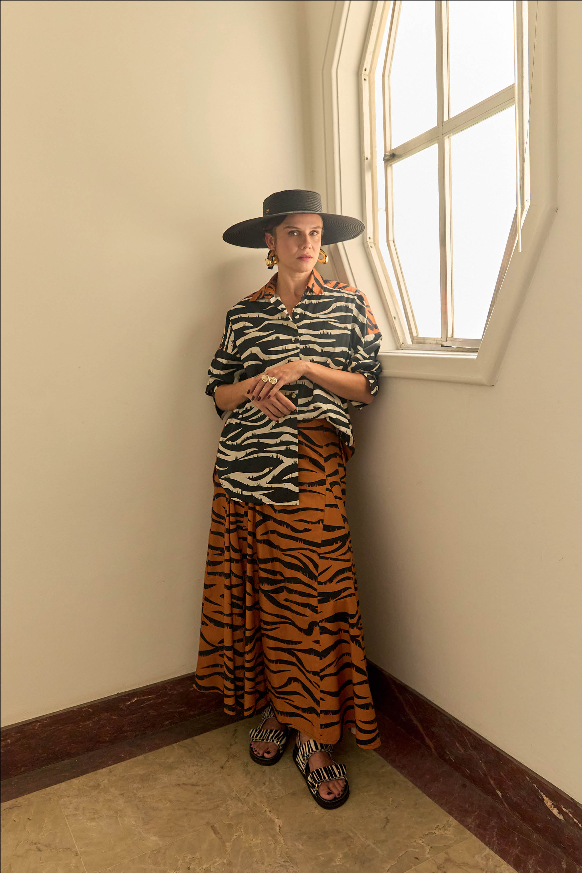 Mulher de pele clara, chap&eacute;u preto de abas largas, camisa de zebra preta e branca, saia longa de tigre laranja e preta, e sand&aacute;lias de zebra, posando em um canto com uma janela ao lado