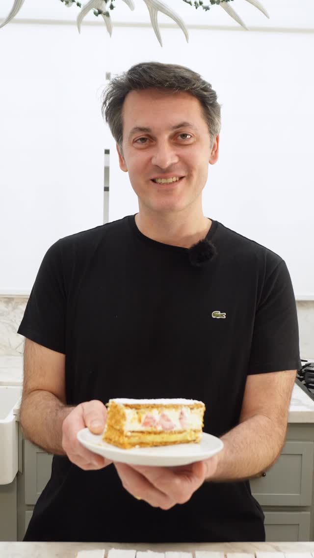 Homem sorridente de camiseta preta, com microfone de lapela, oferece um peda&ccedil;o de bolo mil-folhas com creme e morangos em um prato branco, em uma cozinha clara