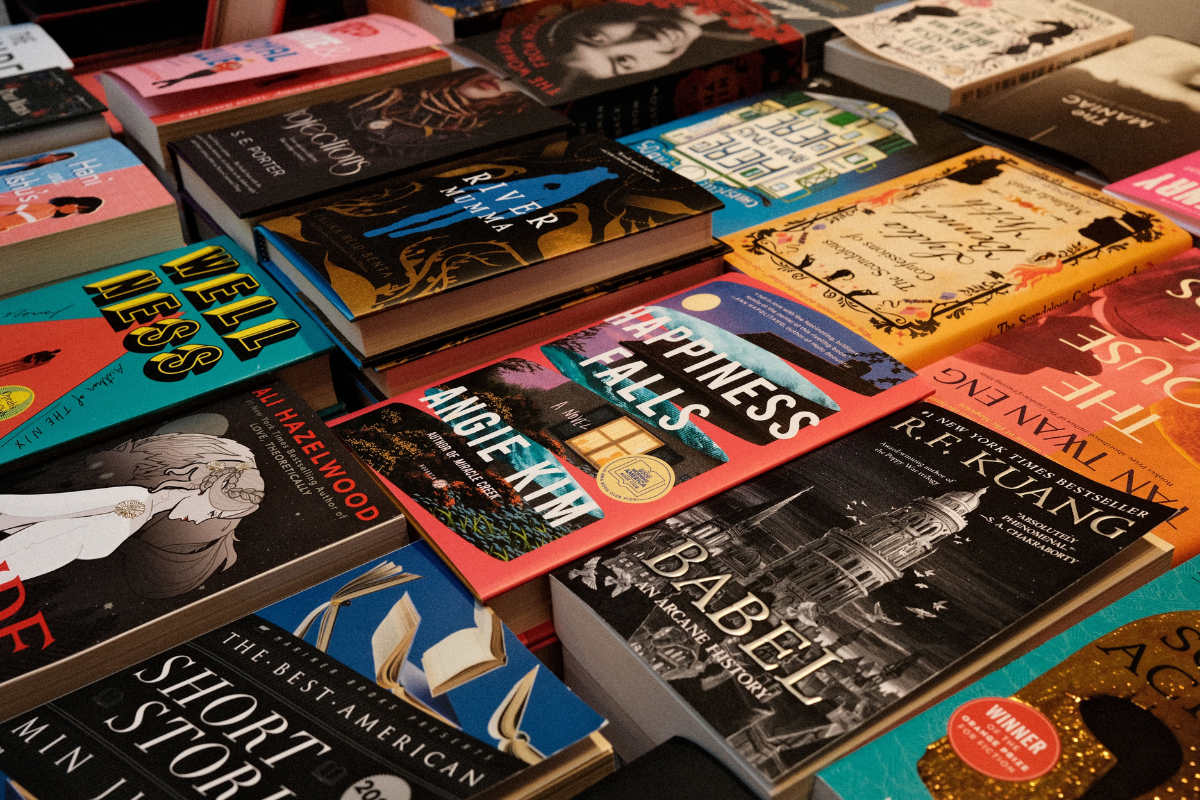 Uma variedade de livros de capa dura e brochura empilhados e dispostos em uma superfície, com títulos como Happiness Falls, Babel e River Mumma visíveis. As capas apresentam ilustrações diversas e coloridas, sugerindo diferentes gêneros literários, como ficção e fantasia. A iluminação destaca os detalhes das capas e a textura dos livros, criando uma atmosfera convidativa para a leitura