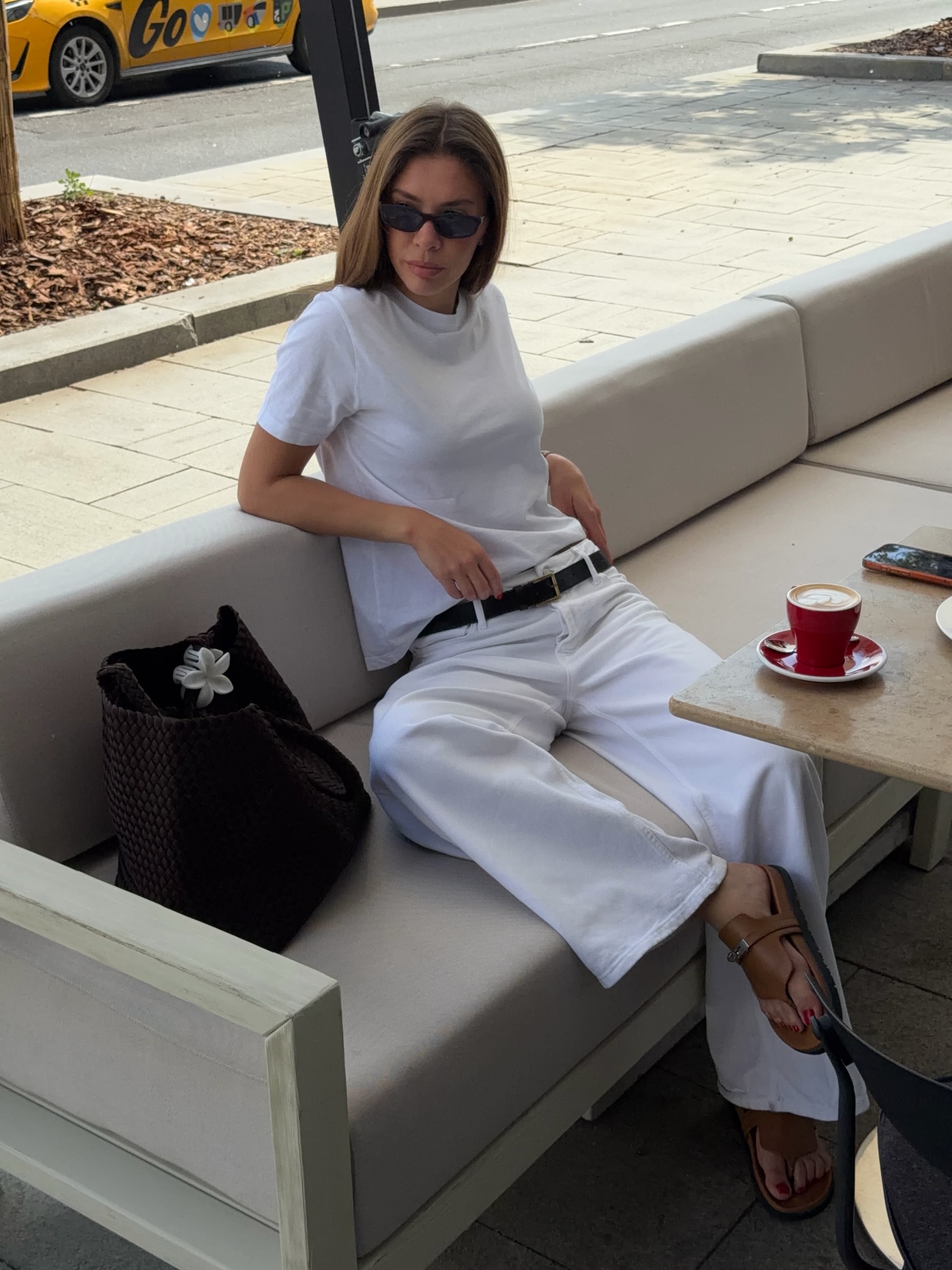 Mulher jovem de &oacute;culos escuros, camiseta branca e cal&ccedil;a branca, sentada relaxadamente em um sof&aacute; bege ao ar livre, com uma x&iacute;cara de caf&eacute; e uma bolsa escura ao lado