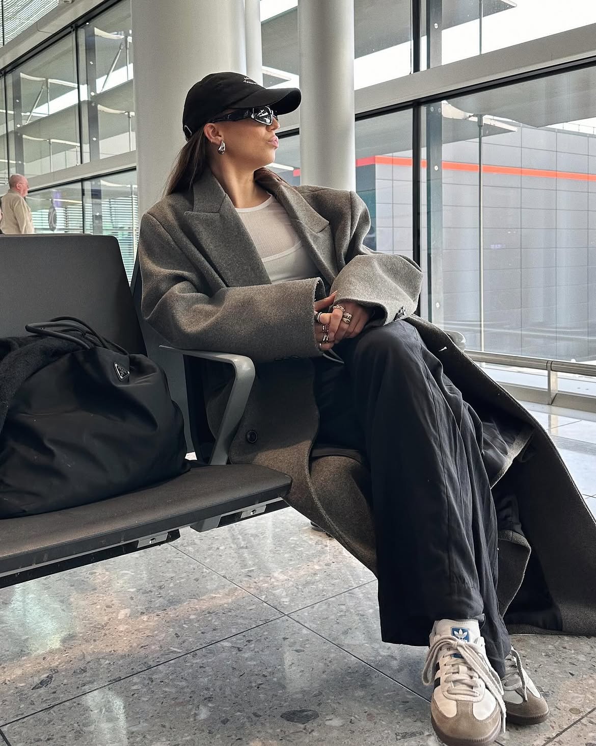 Mulher jovem, de perfil, sentada em banco de aeroporto, usando bon&eacute; preto, &oacute;culos escuros, casaco cinza, camiseta branca, cal&ccedil;a preta e t&ecirc;nis branco com detalhes cinzas. Ela olha para a direita, com uma bolsa preta ao lado. Ao fundo, janelas e um homem desfocado