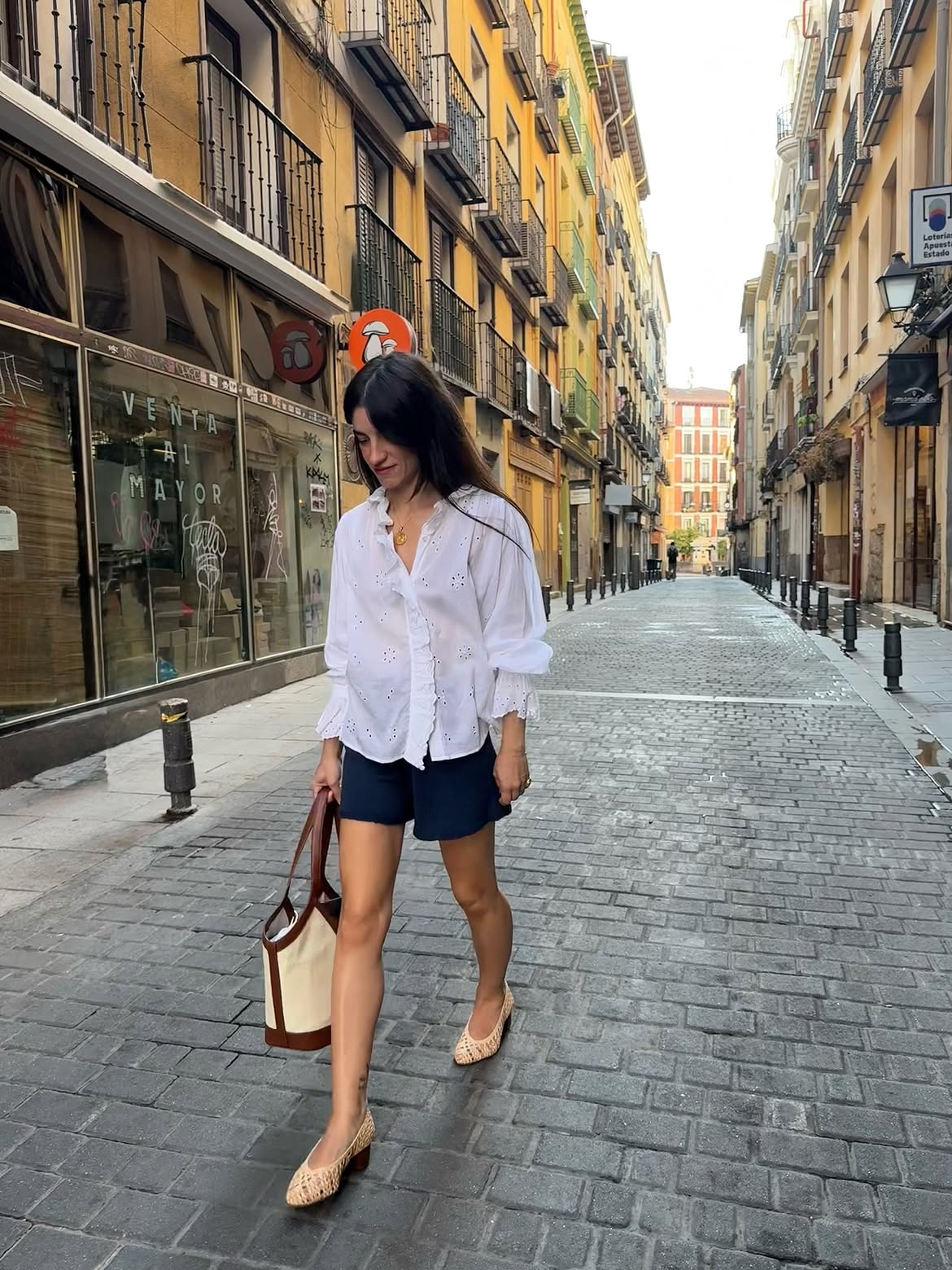 Mulher de cabelos longos e escuros, vestindo blusa branca bordada, shorts azul-marinho e sapatos de salto baixo bege, caminhando em rua estreita de paralelep&iacute;pedos com pr&eacute;dios coloridos e varandas dos dois lados. Ela carrega uma bolsa bege e marrom no bra&ccedil;o esquerdo, olhando para baixo com express&atilde;o pensativa.