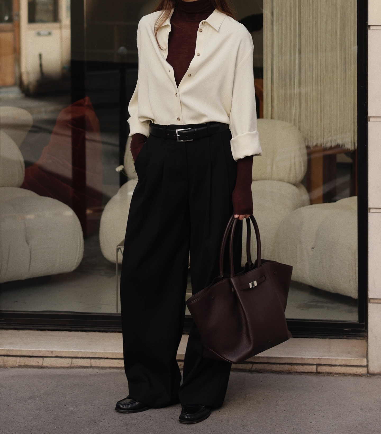 Pessoa de p&eacute;, vestindo camisa bege clara sobre blusa de gola alta vinho, cal&ccedil;a preta larga, cinto preto e sapatos pretos. Segura uma bolsa de couro marrom escura. Ao fundo, uma vitrine reflete o ambiente externo e exibe m&oacute;veis claros no interior