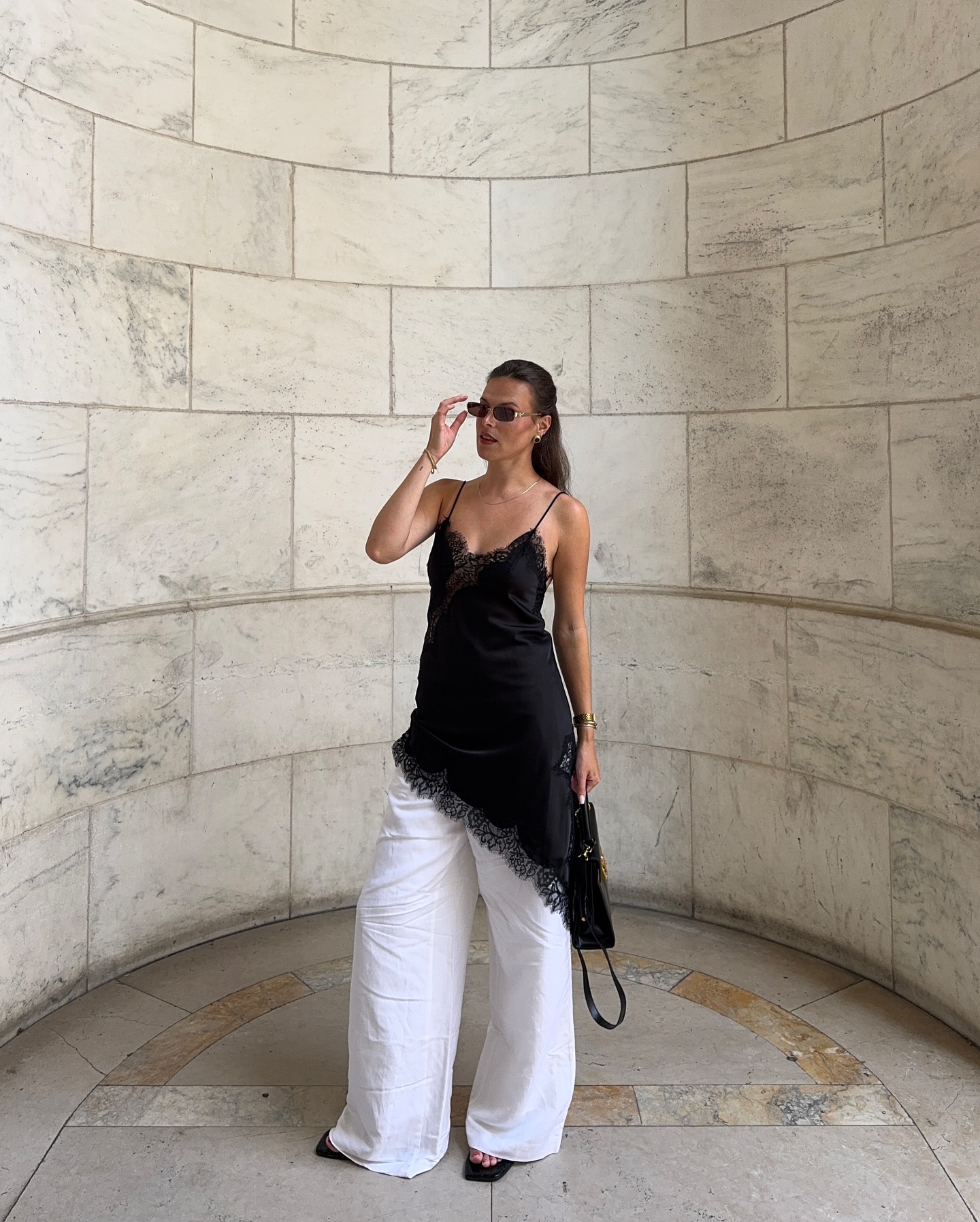 Mulher em p&eacute;, com blusa preta de al&ccedil;as finas e renda, cal&ccedil;a pantalona branca e sand&aacute;lias pretas, ajustando &oacute;culos de sol. Ela segura uma bolsa preta e est&aacute; em um corredor curvo com paredes de m&aacute;rmore claro e piso de pedra