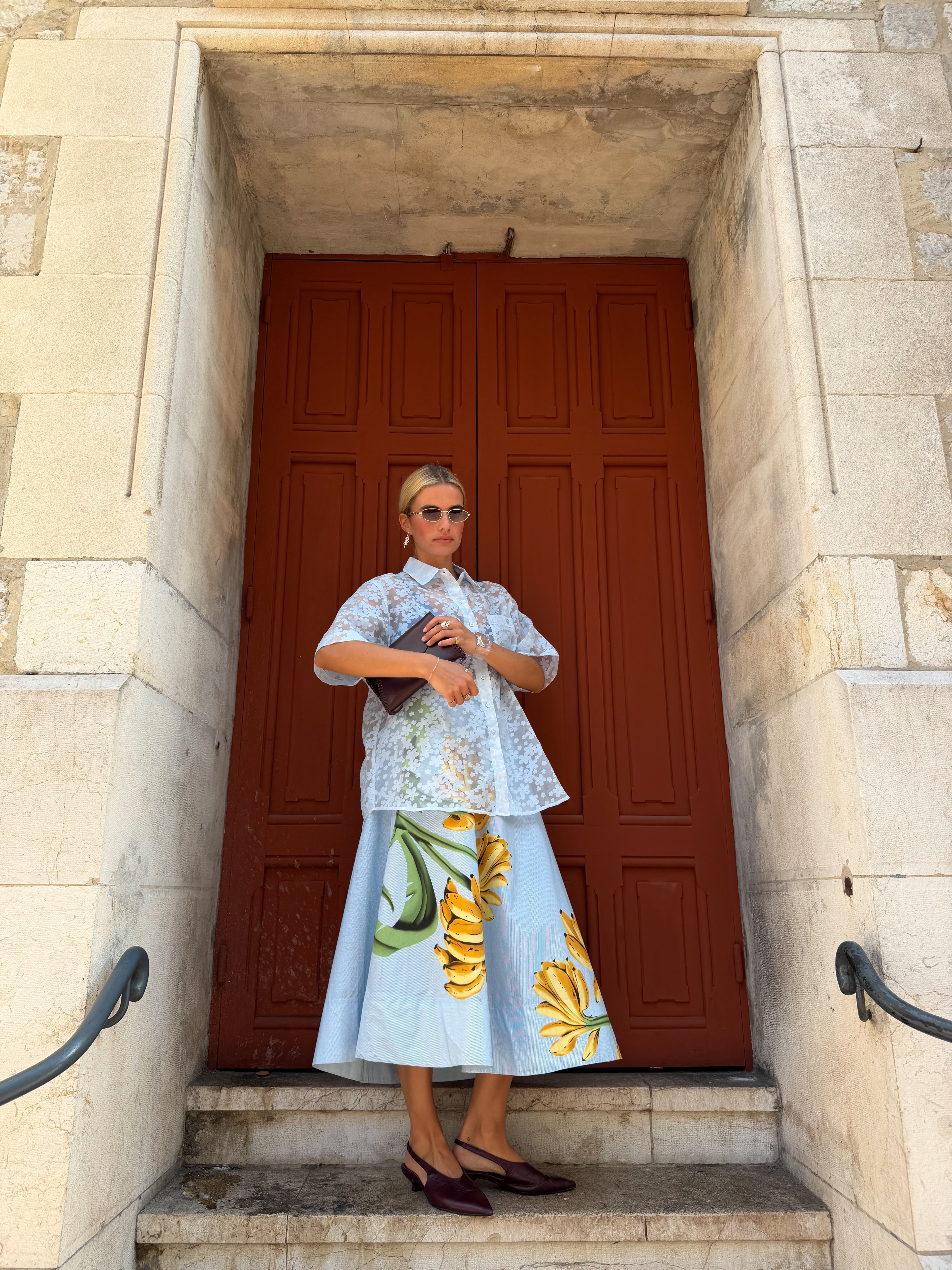 Mulher loira, de &oacute;culos escuros, camisa branca transparente e saia azul com estampa de bananas amarelas, posa em frente a uma porta marrom de madeira, sobre degraus de pedra