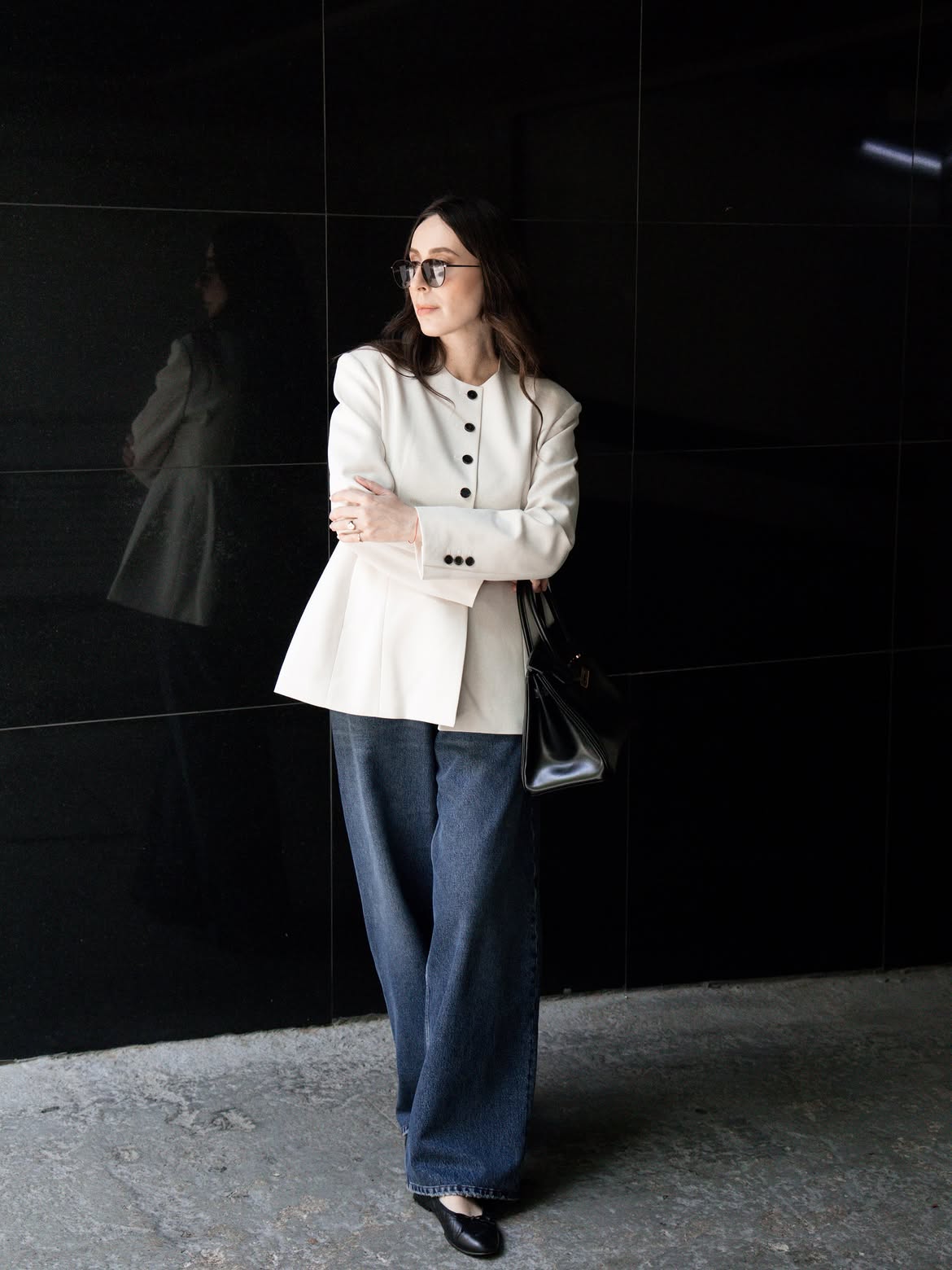 Mulher de cabelos castanhos longos, &oacute;culos escuros, blazer creme com bot&otilde;es pretos, cal&ccedil;a jeans larga e sapatilhas pretas, posando com os bra&ccedil;os cruzados e uma bolsa preta no ombro, em frente a uma parede escura e lisa, com reflexo sutil na lateral
