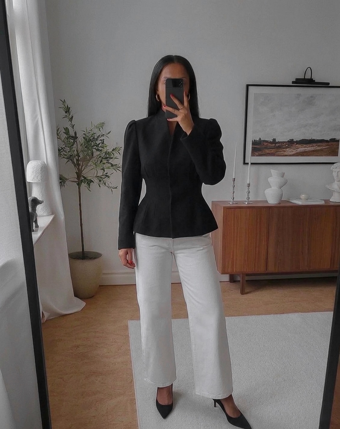 Mulher de cabelos escuros posa para selfie no espelho, vestindo blazer preto com ombreiras e cal&ccedil;a branca larga, sapatos pretos de salto. Ao fundo, planta, c&ocirc;moda de madeira com objetos decorativos e quadro na parede