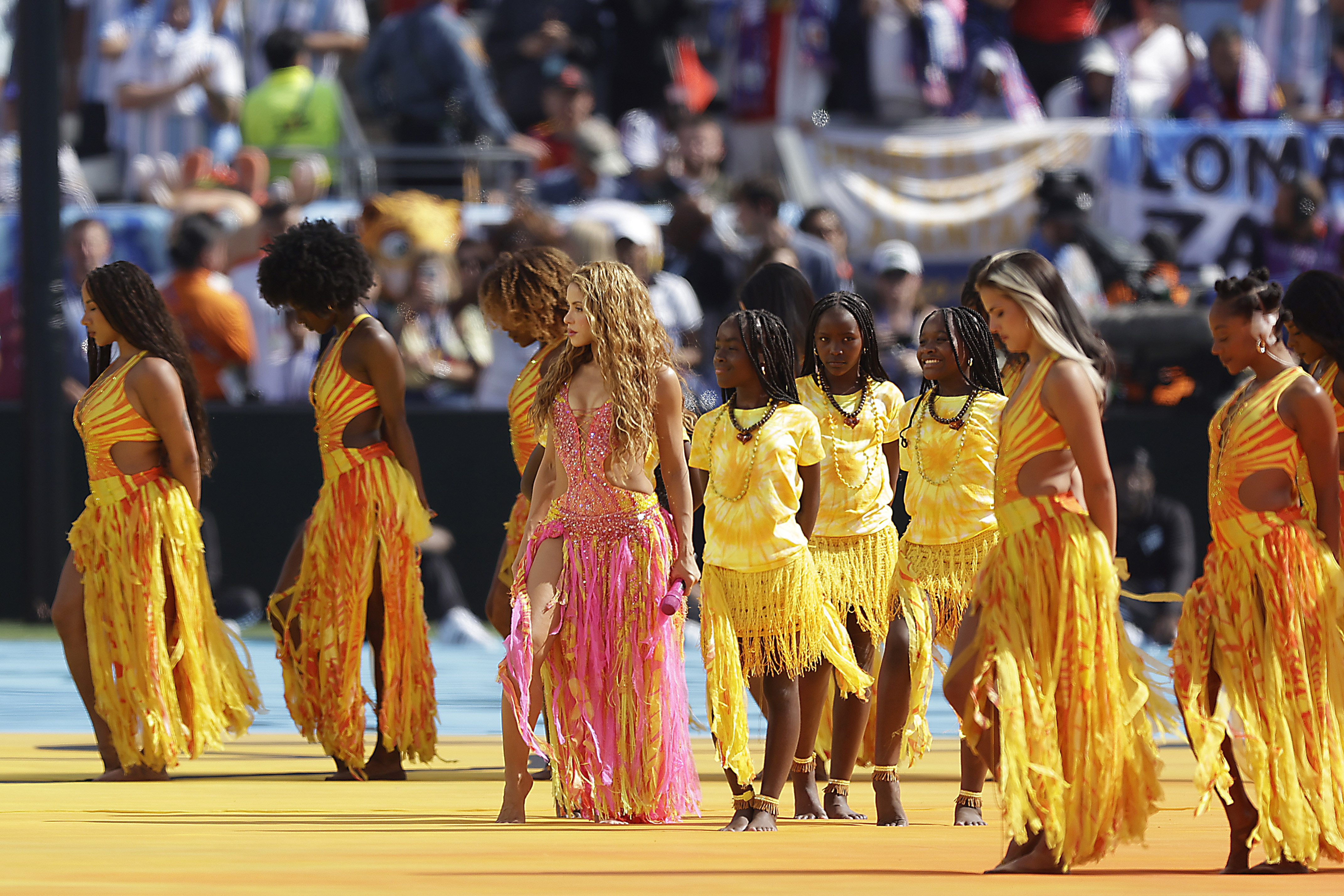 Shakira, de cabelo ondulado e vestido rosa brilhante, caminha descal&ccedil;a com dan&ccedil;arinas em trajes amarelos e laranjas, incluindo crian&ccedil;as, em um palco azul e amarelo. O p&uacute;blico desfocado aparece ao fundo
