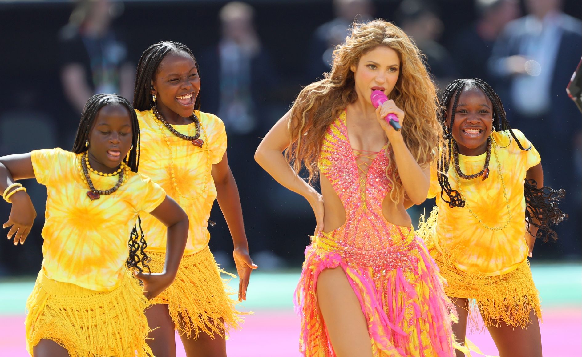 Shakira, com cabelo loiro ondulado e vestido rosa e laranja brilhante, canta em um microfone rosa. Três meninas negras, com tranças e roupas amarelas tie-dye com franjas, dançam ao lado dela, sorrindo e gesticulando com os braços. O fundo está desfocado, com tons escuros e claros