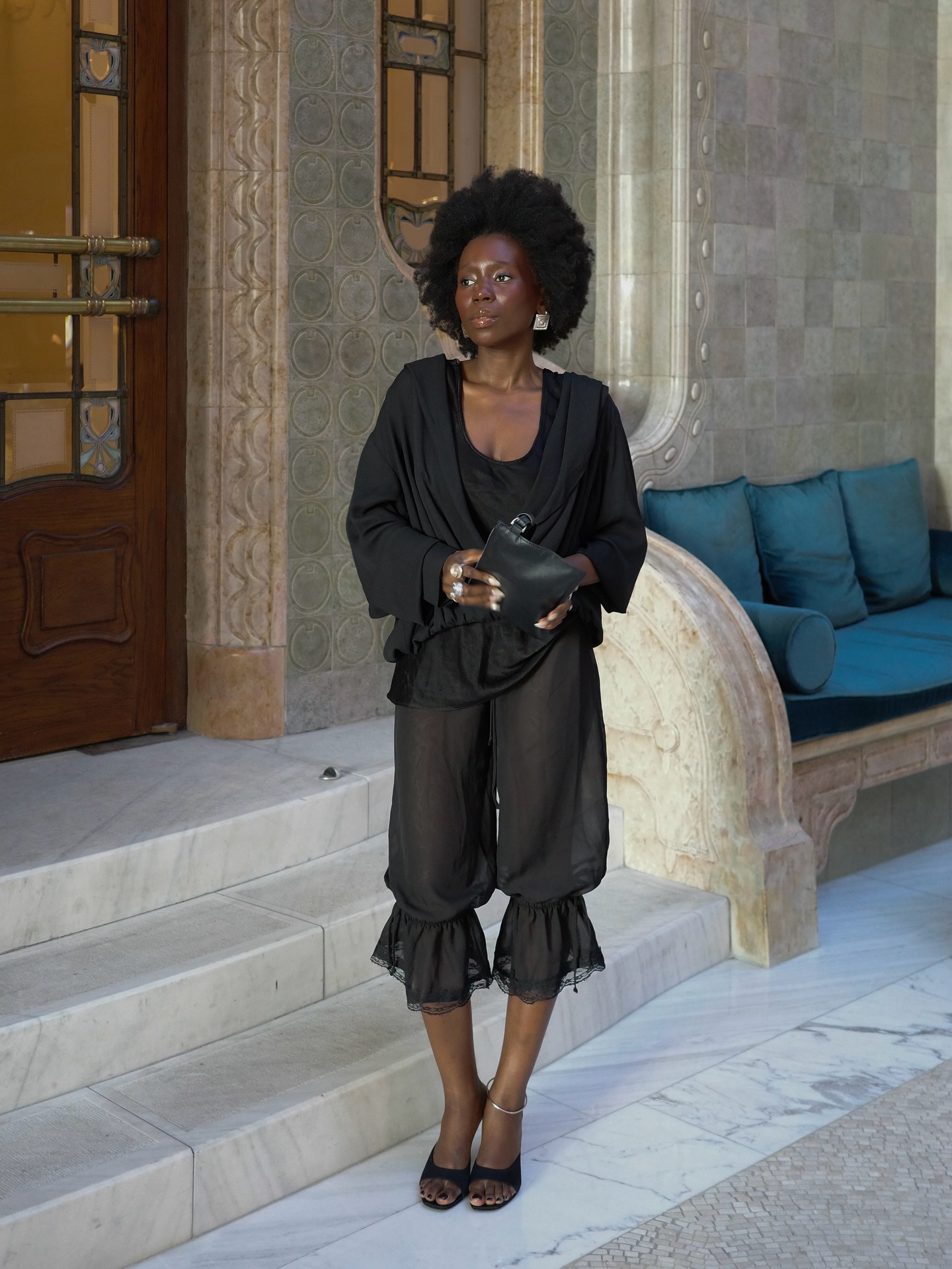 Mulher negra de cabelo afro, vestindo blusa preta solta, cal&ccedil;a preta transparente com babados e sand&aacute;lias de salto, segurando uma bolsa preta. Ela est&aacute; em p&eacute; em uma escadaria de m&aacute;rmore, com uma porta de madeira e uma parede de azulejos ao fundo, e um sof&aacute; azul &agrave; direita
