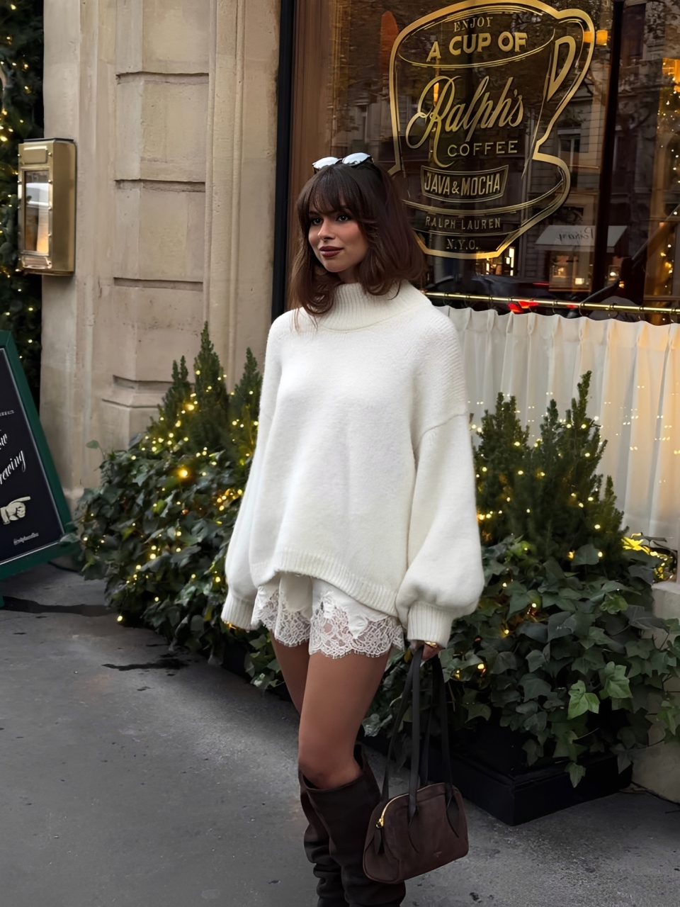 Mulher jovem com cabelo castanho e franja, usando su&eacute;ter branco de gola alta, shorts de renda branca e botas marrons, segurando uma bolsa marrom. Ela est&aacute; em frente a uma vitrine com a inscri&ccedil;&atilde;o Ralphs Coffee e arbustos decorados com luzes