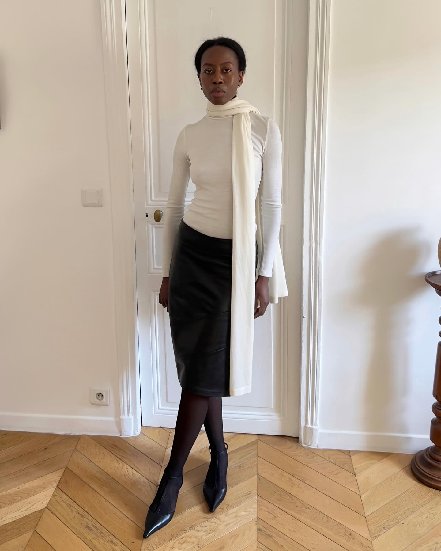 Mulher negra de p&eacute;, com express&atilde;o s&eacute;ria, vestindo blusa branca de gola alta, saia l&aacute;pis preta de couro, meias-cal&ccedil;as pretas e sapatos de bico fino. Um len&ccedil;o branco comprido pende do pesco&ccedil;o, sobre a blusa. Ela est&aacute; em um ambiente interno com porta branca e piso de madeira em padr&atilde;o chevron