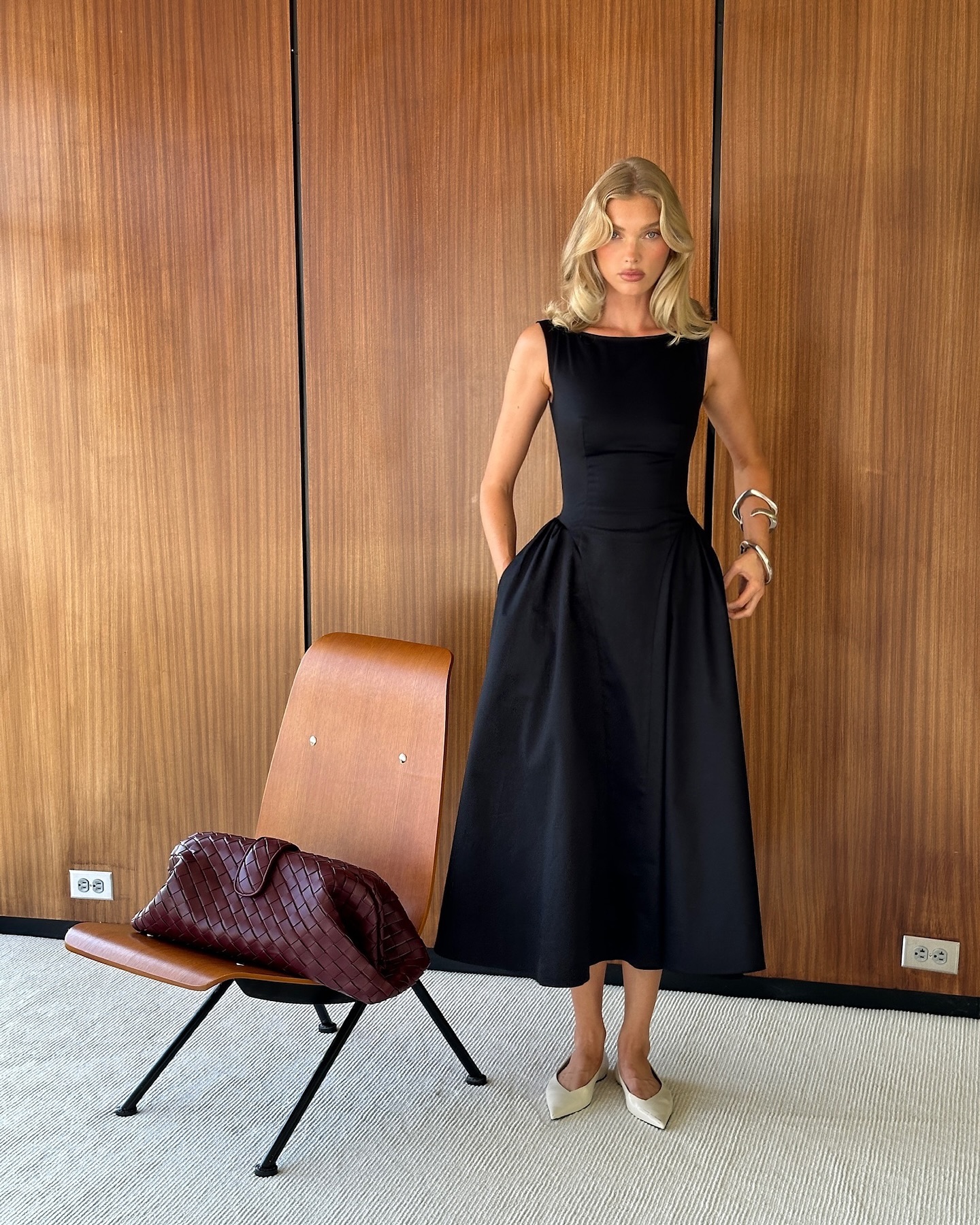 Mulher loira de cabelo ondulado, vestindo um elegante vestido preto sem mangas e sapatos brancos, posa com as m&atilde;os nos bolsos. Ela usa pulseiras prateadas no bra&ccedil;o esquerdo. Ao lado, uma cadeira de madeira com uma bolsa marrom escura tran&ccedil;ada. O fundo &eacute; uma parede de madeira com detalhes verticais e um tapete claro.