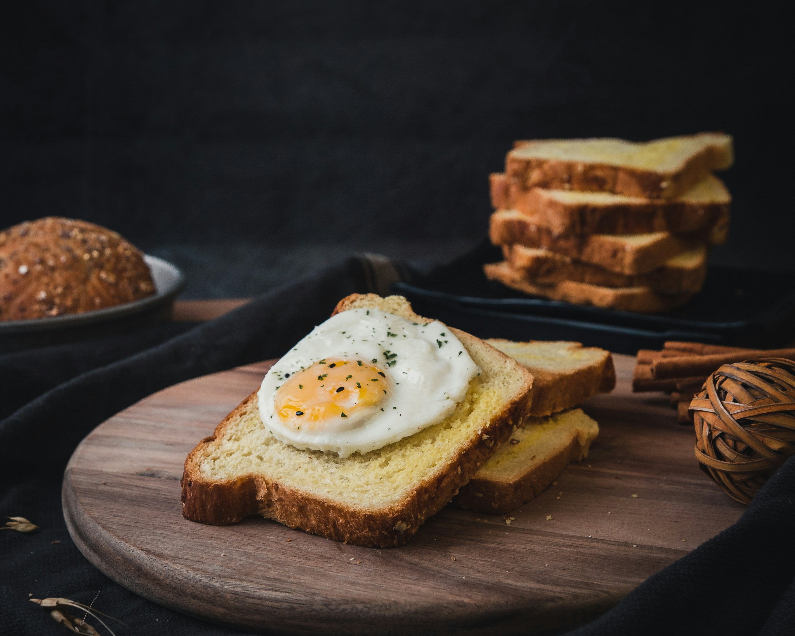 Fatia de p&atilde;o de forma com ovo frito e gema mole, polvilhado com temperos, sobre uma t&aacute;bua de madeira. Ao fundo, p&atilde;es e torradas empilhadas, com canela em pau e um objeto decorativo de vime, em um ambiente escuro