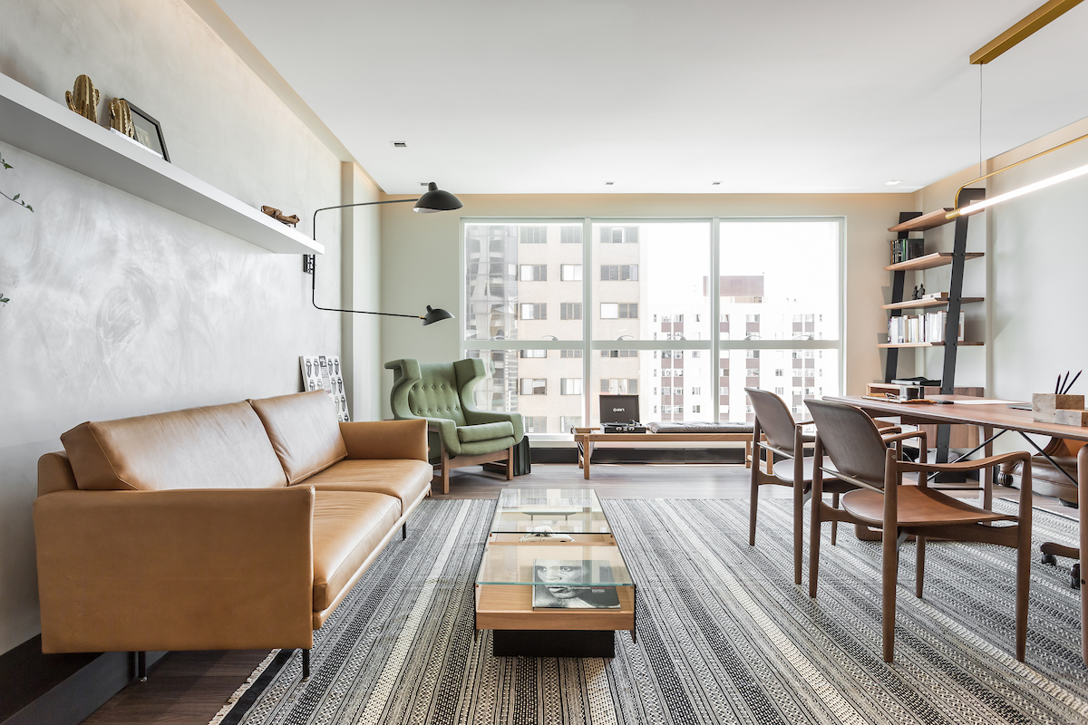 Sala de estar e escrit&oacute;rio integrados com sof&aacute; de couro caramelo, poltrona verde, mesa de centro de madeira e vidro, e mesa de trabalho com duas cadeiras de madeira e couro. O ambiente tem um tapete listrado, prateleiras com livros e uma grande janela com vista para pr&eacute;dios
