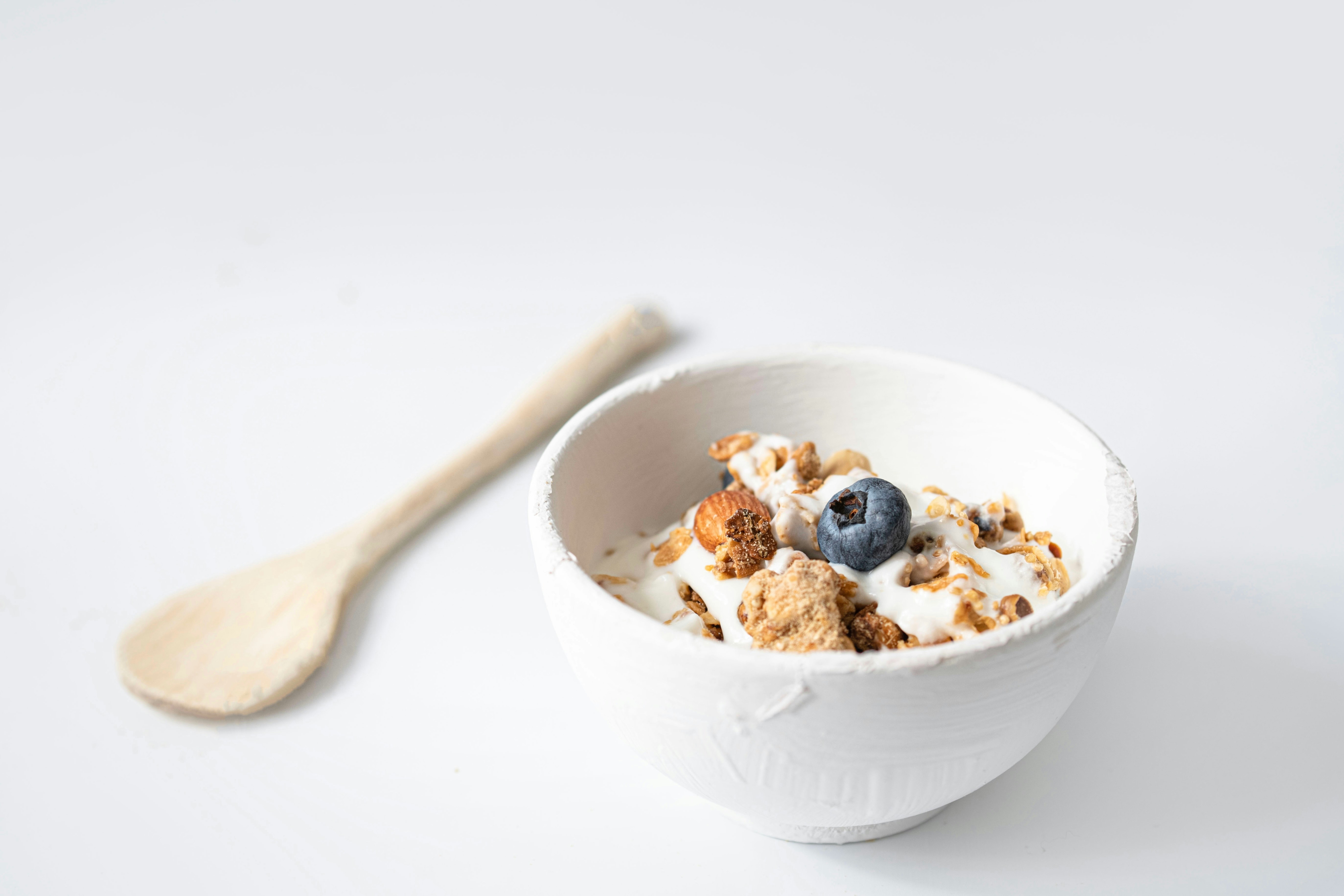 Tigela branca com iogurte, granola e uma amora no centro, ao lado de uma colher de madeira clara sobre fundo branco