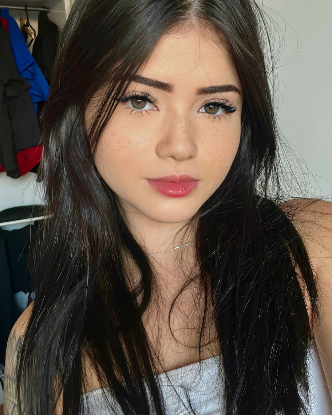 Jovem mulher de cabelos escuros e longos, com sardas no rosto, olhando diretamente para a c&acirc;mera. Ela usa um top branco e um colar prateado fino. Ao fundo, roupas penduradas em um cabideiro.