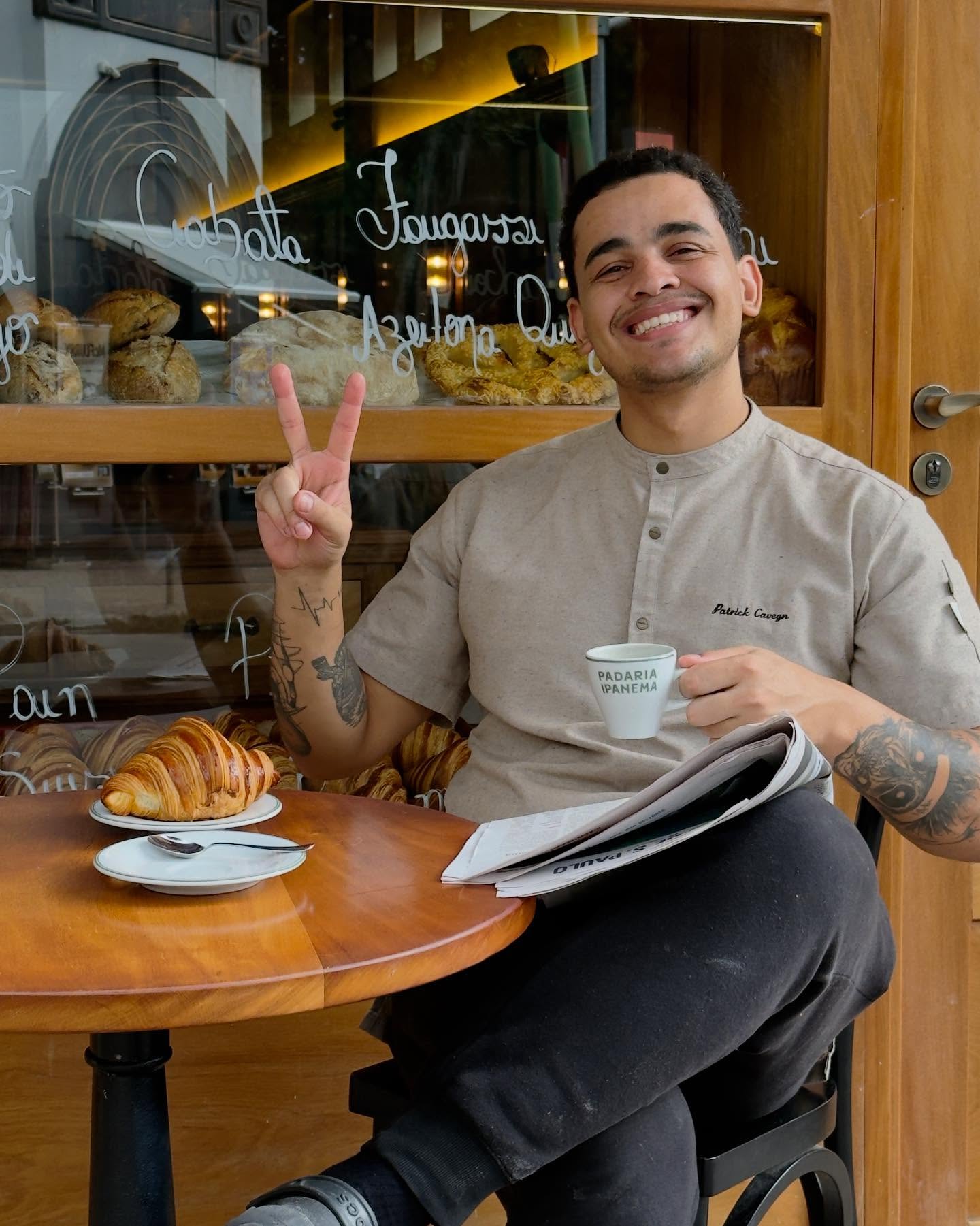 Homem jovem sorridente, de camisa bege e bra&ccedil;os tatuados, sentado em mesa de madeira com croissant, jornal e x&iacute;cara Padaria Ipanema, fazendo sinal de paz com a m&atilde;o esquerda. Ao fundo, vitrine de padaria com p&atilde;es e letreiros brancos Ciabatta, Fougasse, Azeitona