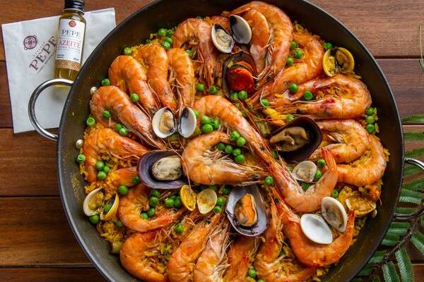 Paella de frutos do mar em uma panela grande, com camarões, mexilhões e vôngoles sobre arroz amarelo e ervilhas verdes. Ao lado, uma garrafa de azeite e um guardanapo branco com a marca Pepe.