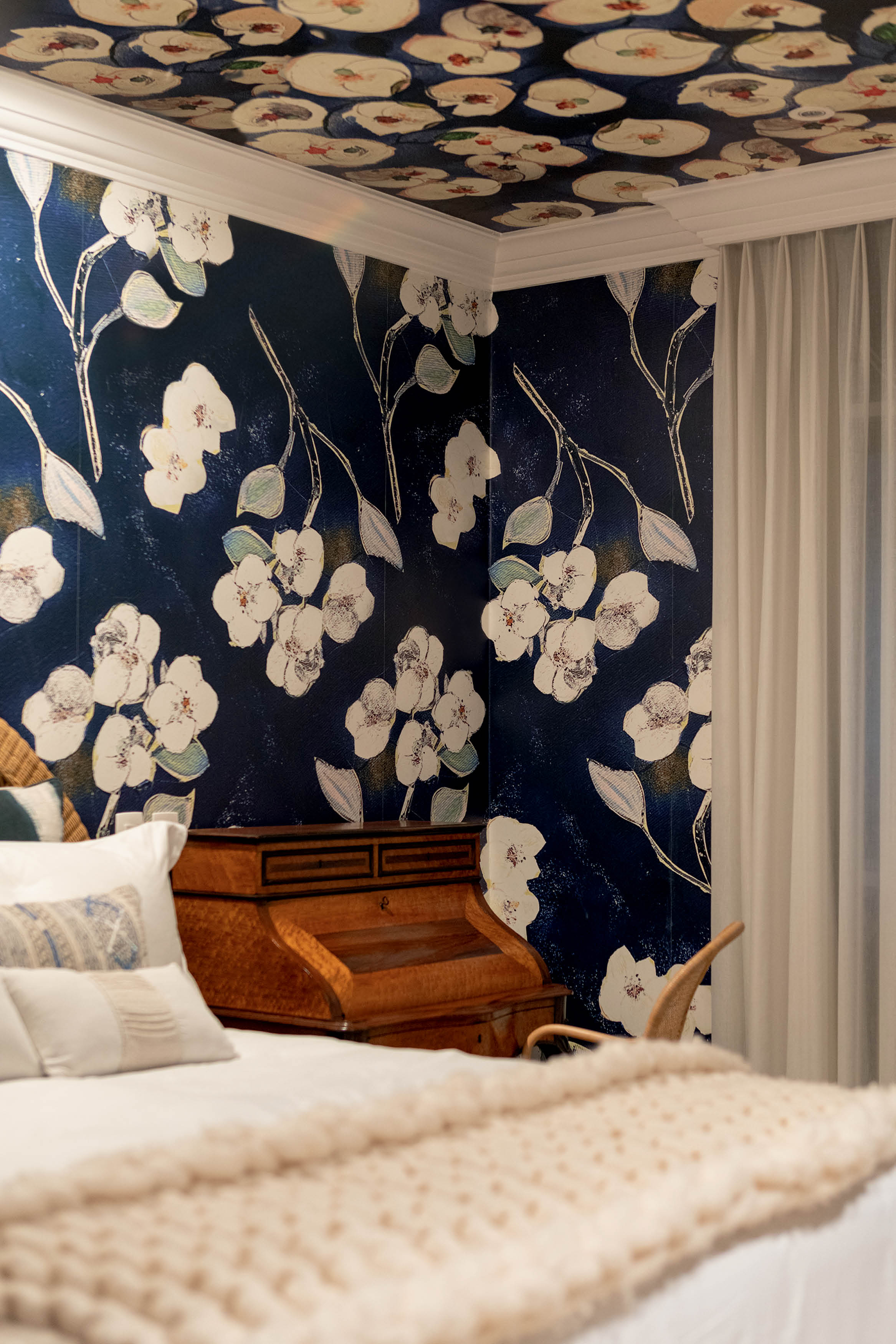 Quarto com papel de parede azul-marinho estampado com flores brancas e folhas claras, combinando com o teto. Uma cama com len&ccedil;&oacute;is brancos e manta bege, e uma escrivaninha de madeira escura com detalhes em marchetaria ao lado. Cortina branca transl&uacute;cida na janela.