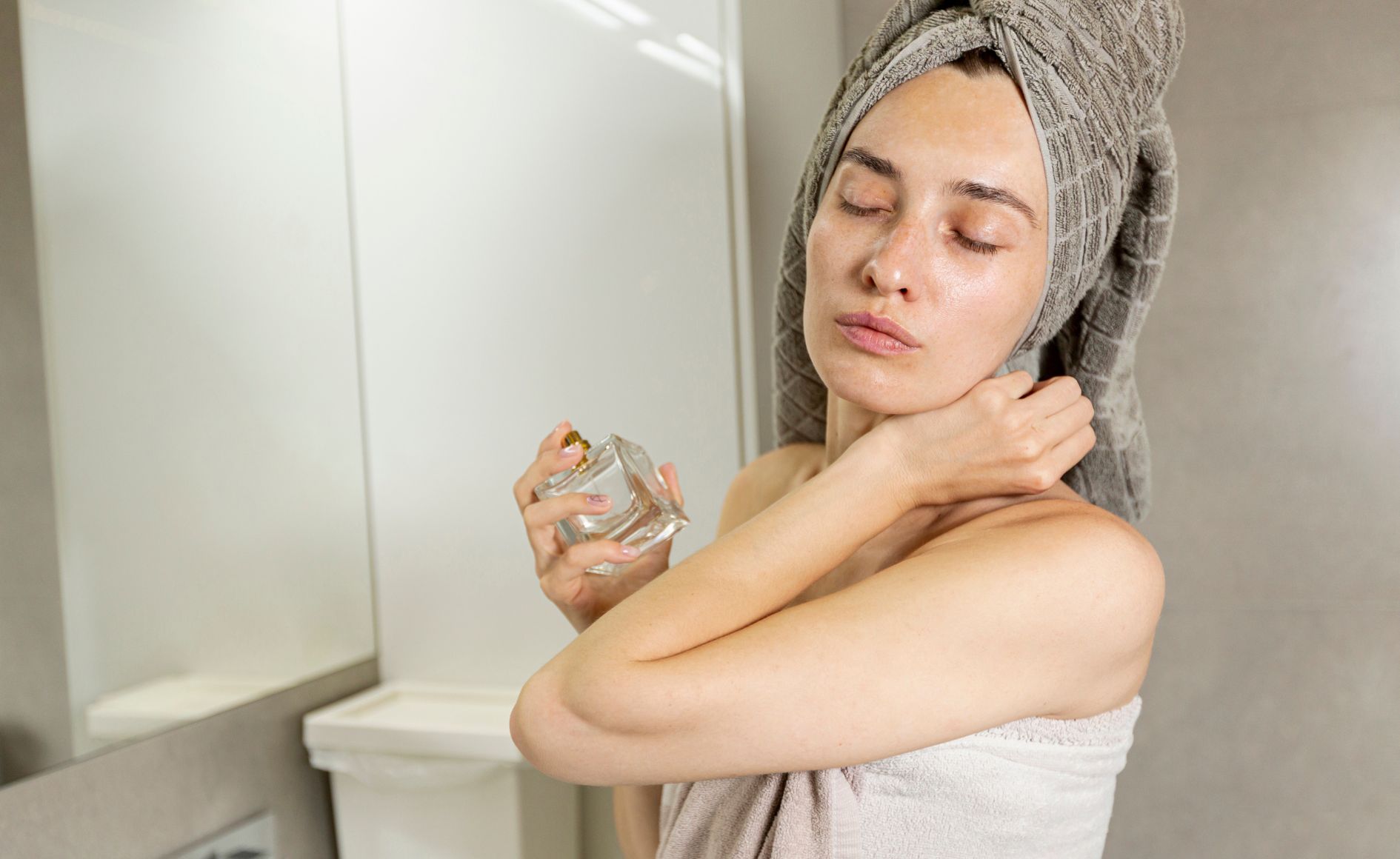Mulher de olhos fechados, com toalha cinza na cabeça e toalha branca no corpo, aplicando perfume no pescoço em um banheiro claro