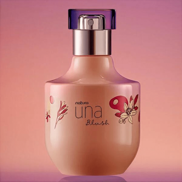 Frasco de perfume Natura Una Blush, cor p&ecirc;ssego, com tampa roxa transl&uacute;cida e borrifador prateado. O frasco tem desenhos florais em tons de rosa e vermelho, e est&aacute; sobre uma superf&iacute;cie roxa, com fundo rosa claro
