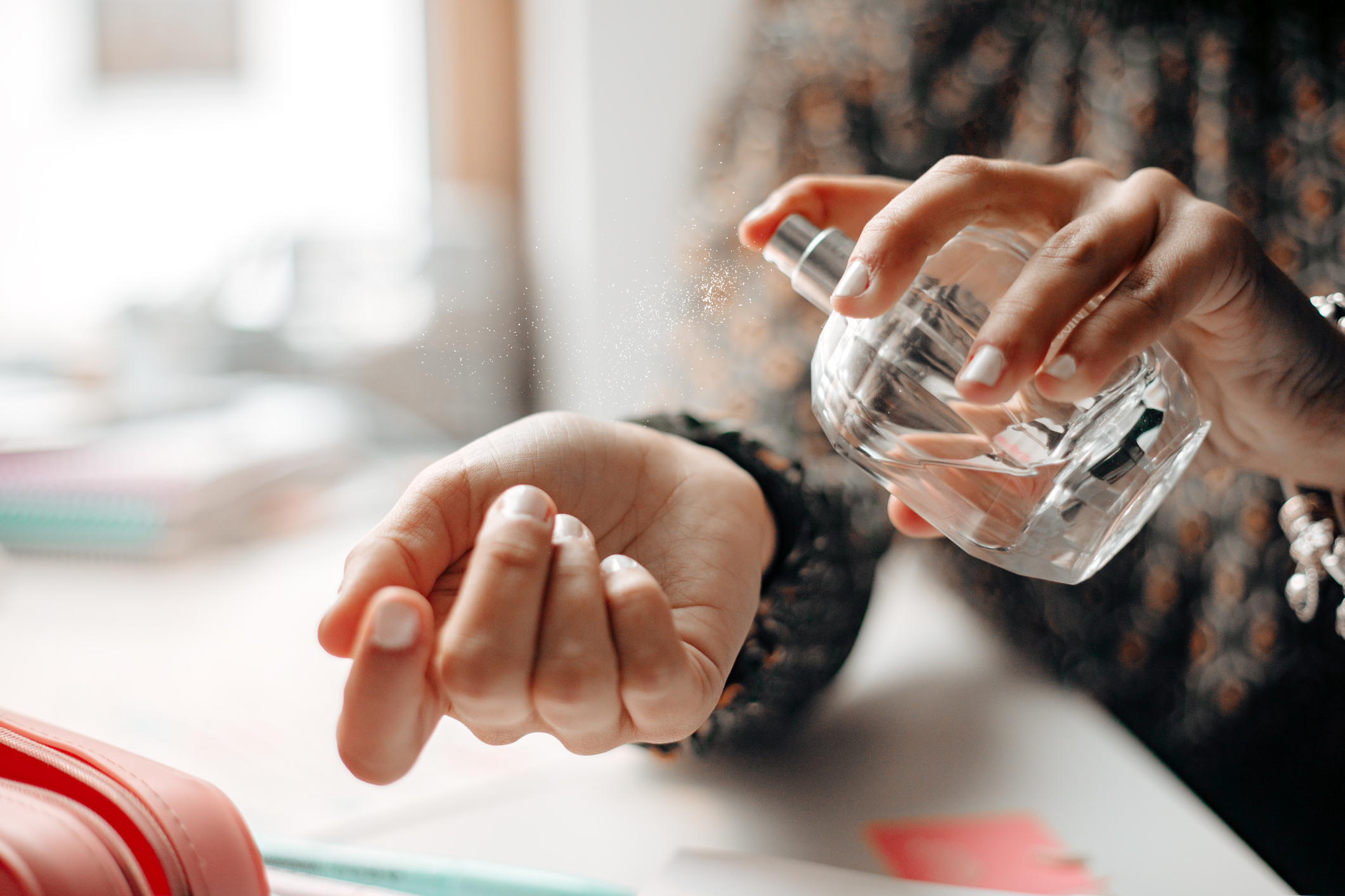 Pessoa com unhas brancas borrifando perfume de um frasco transparente no pulso esquerdo, com gotículas visíveis no ar