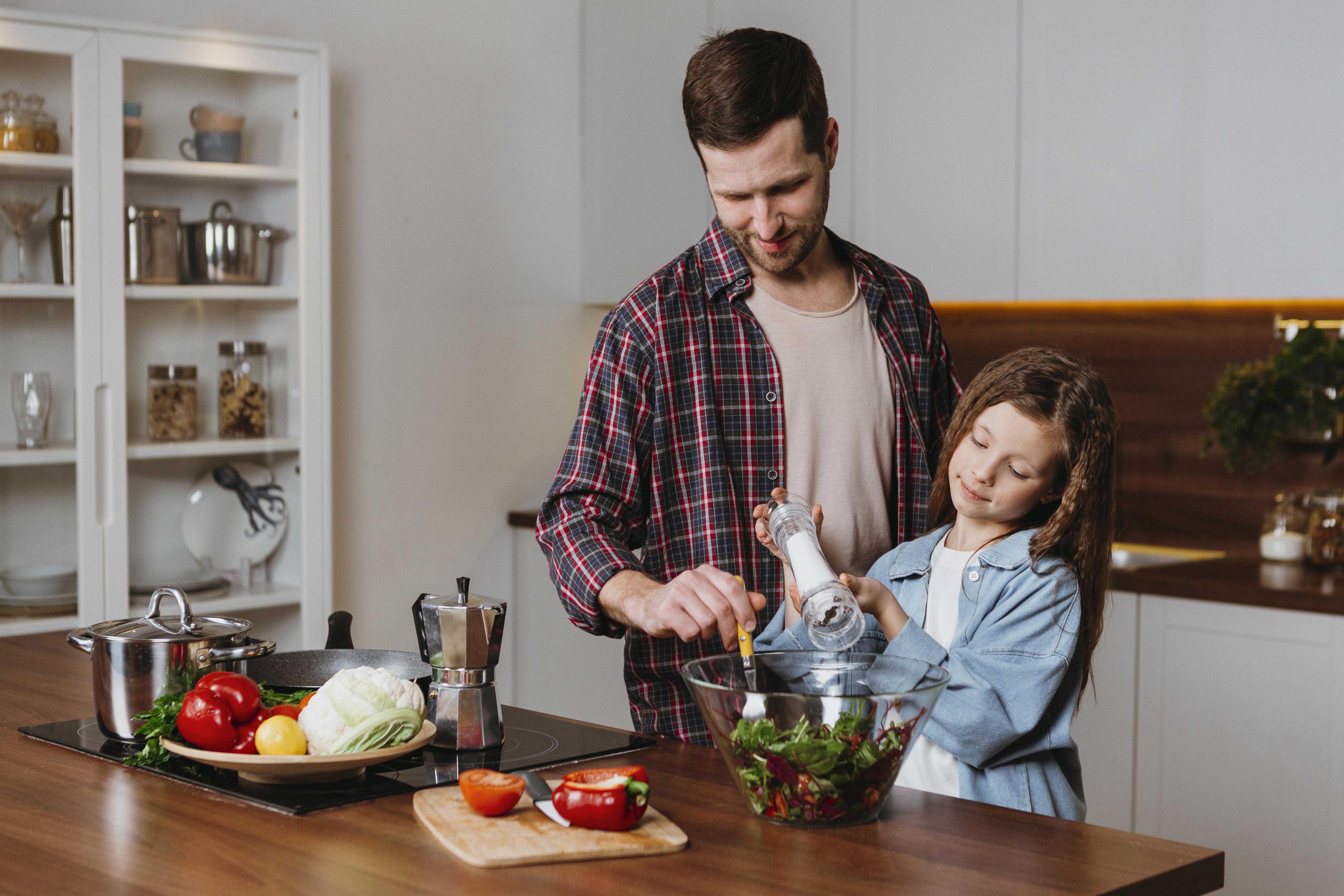 Pai e filha em uma cozinha moderna. O homem, de camisa xadrez, sorri enquanto a menina, de cabelos longos e jaqueta jeans, tempera uma salada com um moedor de sal. Sobre o balc&atilde;o de madeira, h&aacute; legumes frescos, panelas e uma cafeteira.