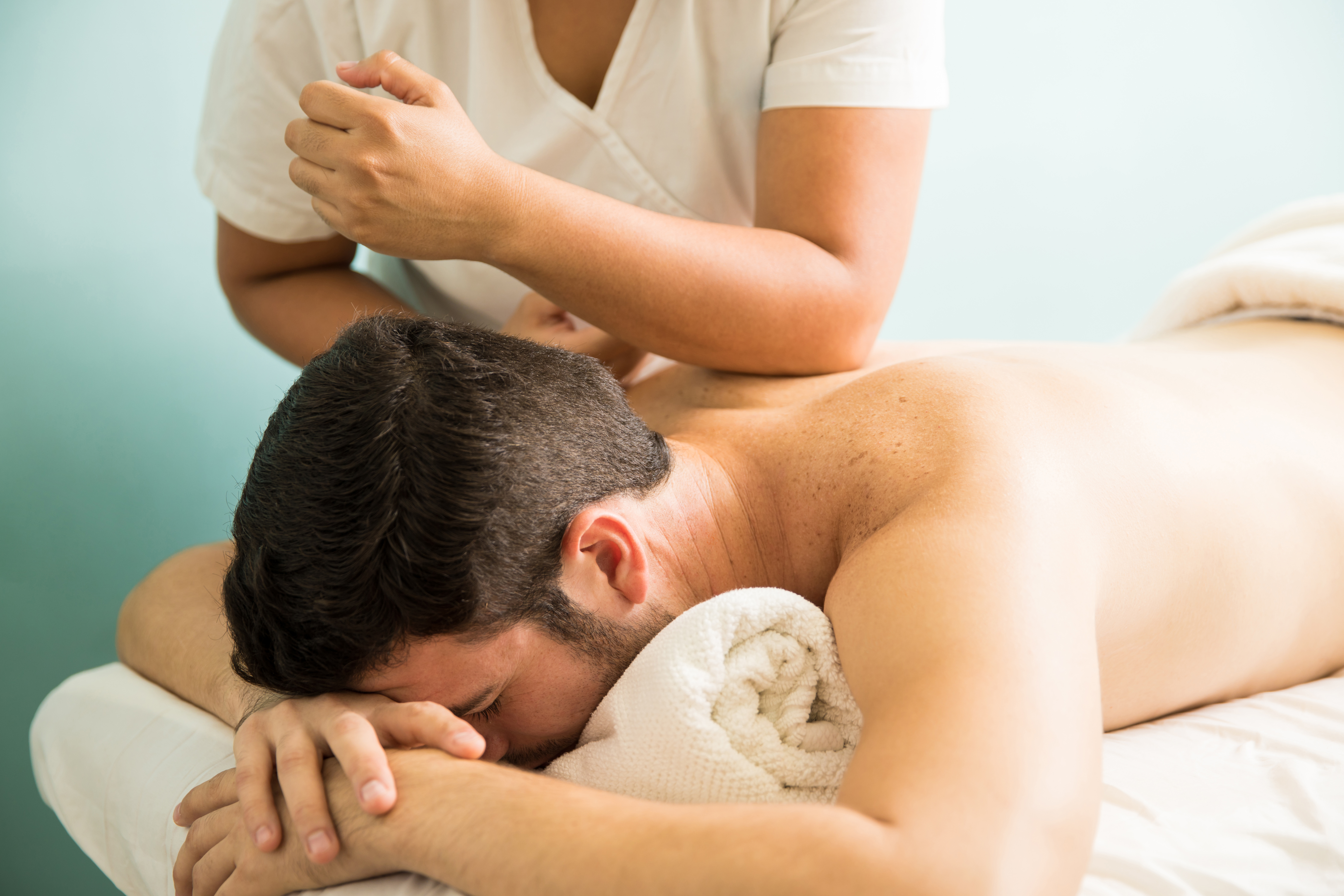 Homem deitado de bru&ccedil;os em uma maca, recebendo massagem nas costas de uma terapeuta com uniforme branco. Ele tem barba e cabelo escuro, com o rosto apoiado em uma toalha branca. A terapeuta usa o cotovelo para massagear a regi&atilde;o do ombro do homem, que parece relaxado. O fundo &eacute; azul claro