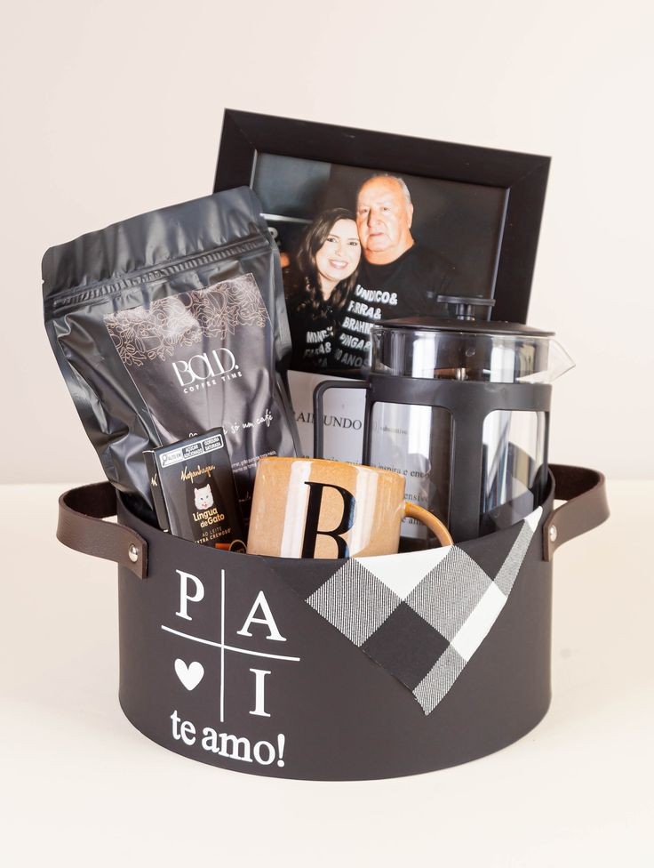 Cesta de presente preta com al&ccedil;as de couro, decorada com PAI te amo! e um xadrez preto e branco. Cont&eacute;m uma foto de um casal sorrindo, um pacote de caf&eacute; Bold, uma prensa francesa e uma caneca com a letra B