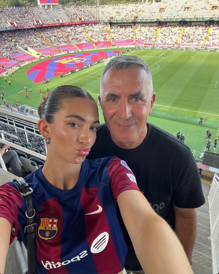 Mulher jovem e homem sorrindo para uma selfie no est&aacute;dio de futebol Camp Nou, com a arquibancada lotada e o campo ao fundo. Ela veste camisa do Barcelona e faz bico, ele usa camiseta preta