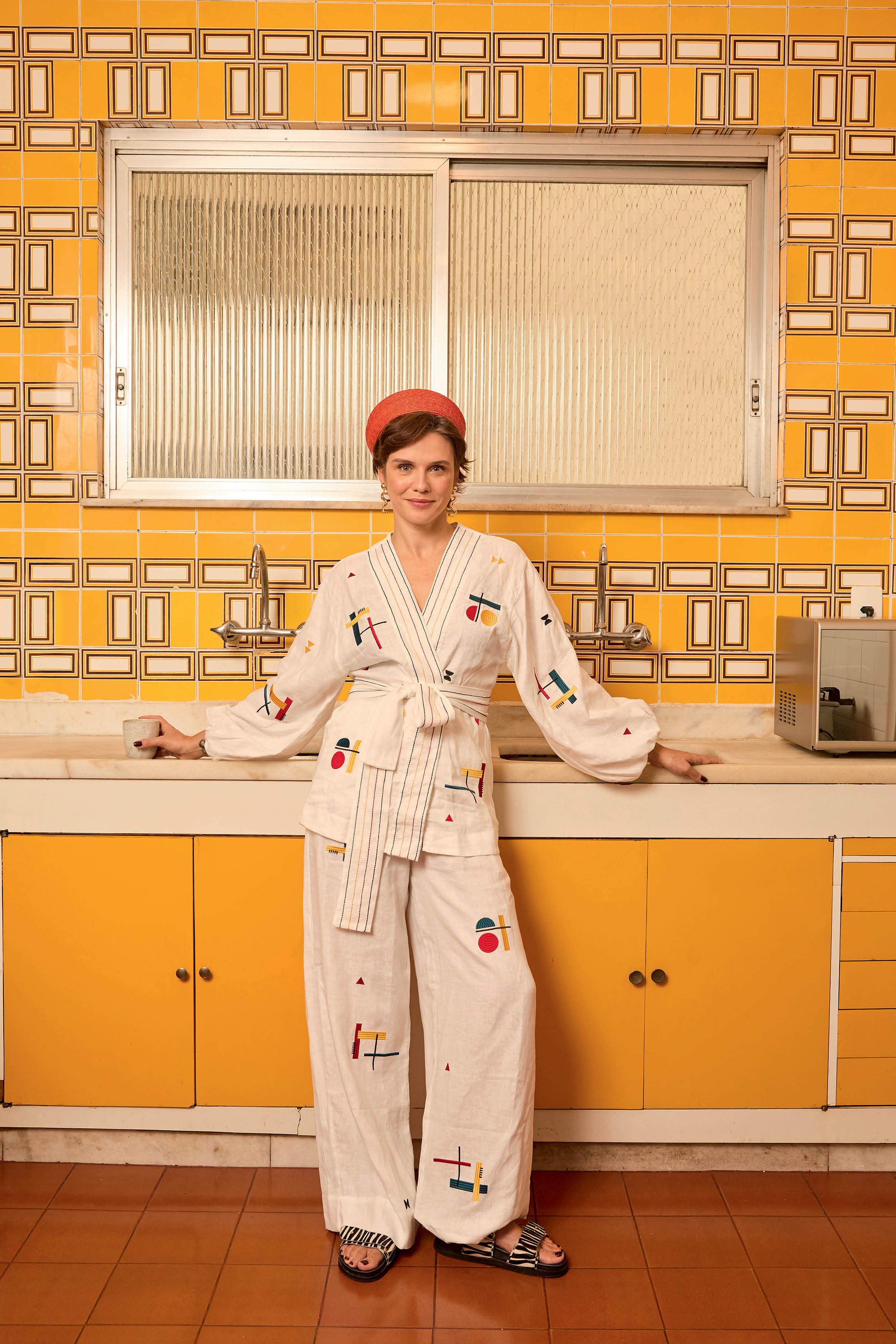 Mulher de cabelo curto e ruivo, usando faixa vermelha na cabe&ccedil;a, brincos dourados e conjunto branco com bordados coloridos, posa sorrindo em uma cozinha com azulejos amarelos e arm&aacute;rios laranjas