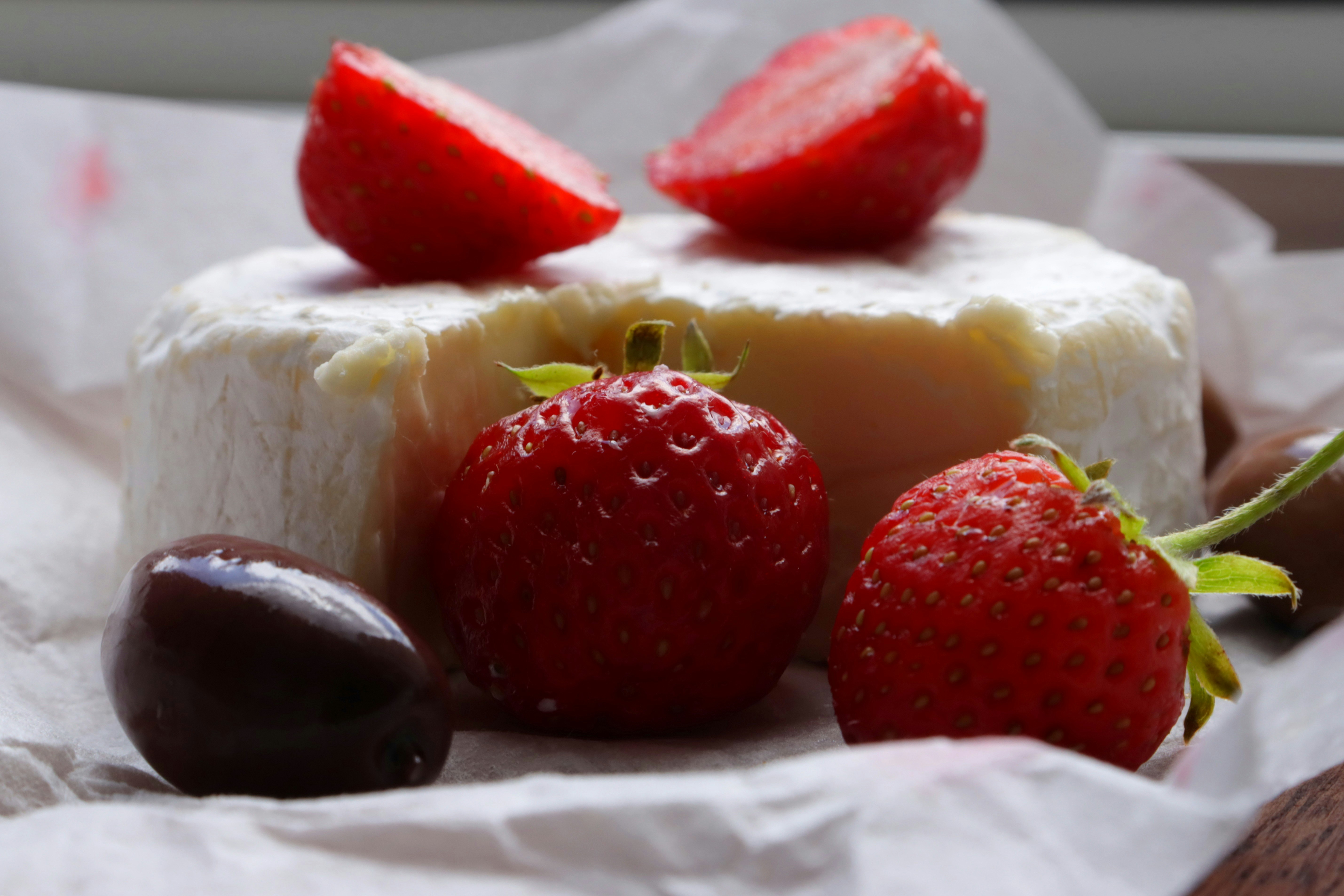 Queijo brie redondo com casca branca, duas metades de morango em cima, um morango inteiro &agrave; frente e uma azeitona preta brilhante &agrave; esquerda, sobre papel amassado