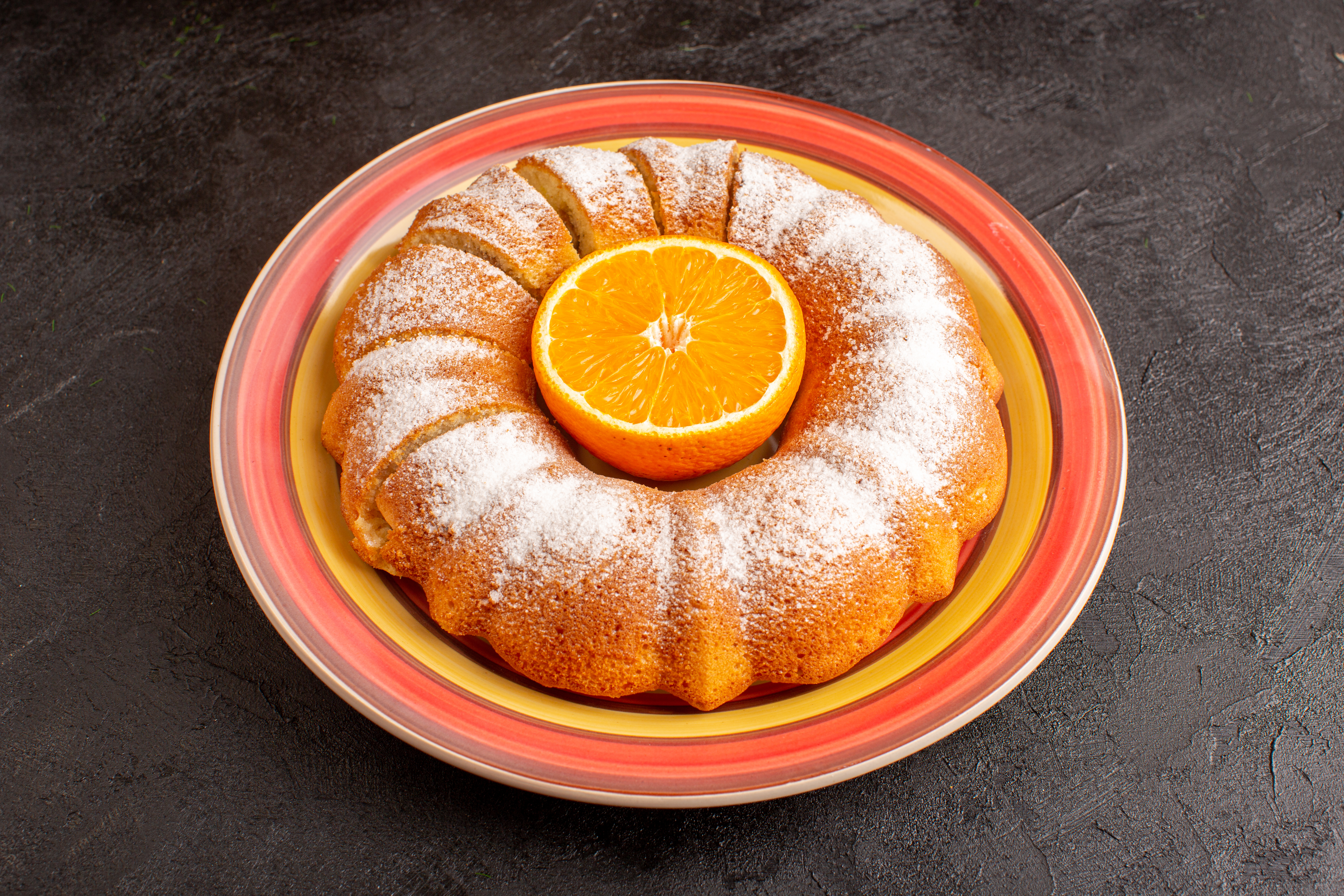 Bolo de laranja em formato de anel, com açúcar de confeiteiro polvilhado e uma metade de laranja no centro, servido em um prato colorido com bordas vermelhas e amarelas, sobre uma superfície escura