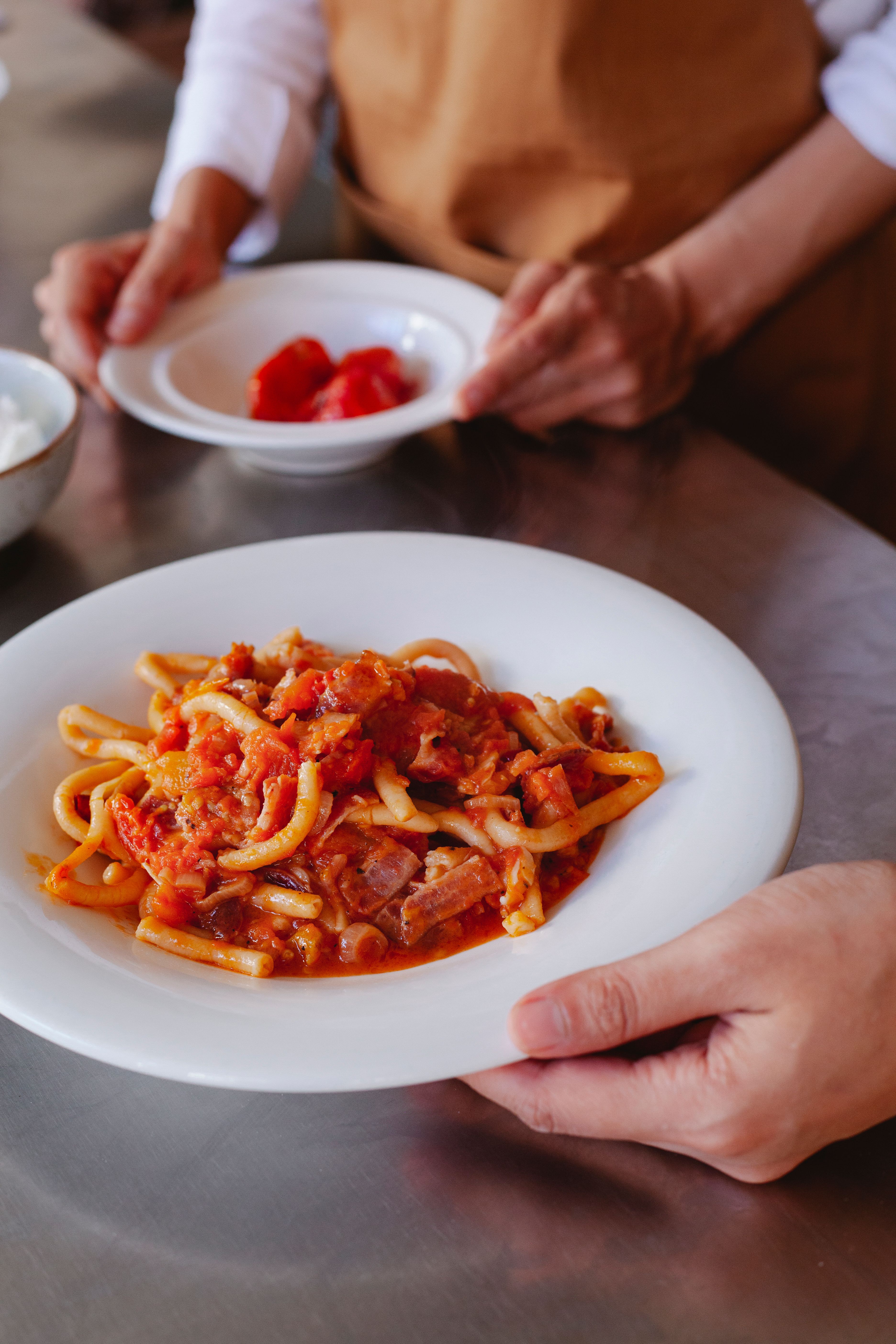 Duas pessoas em uma cozinha, uma delas segurando um prato branco com macarr&atilde;o tipo bucatini e molho de tomate com peda&ccedil;os de carne, enquanto a outra segura uma tigela com tomates picados ao fundo