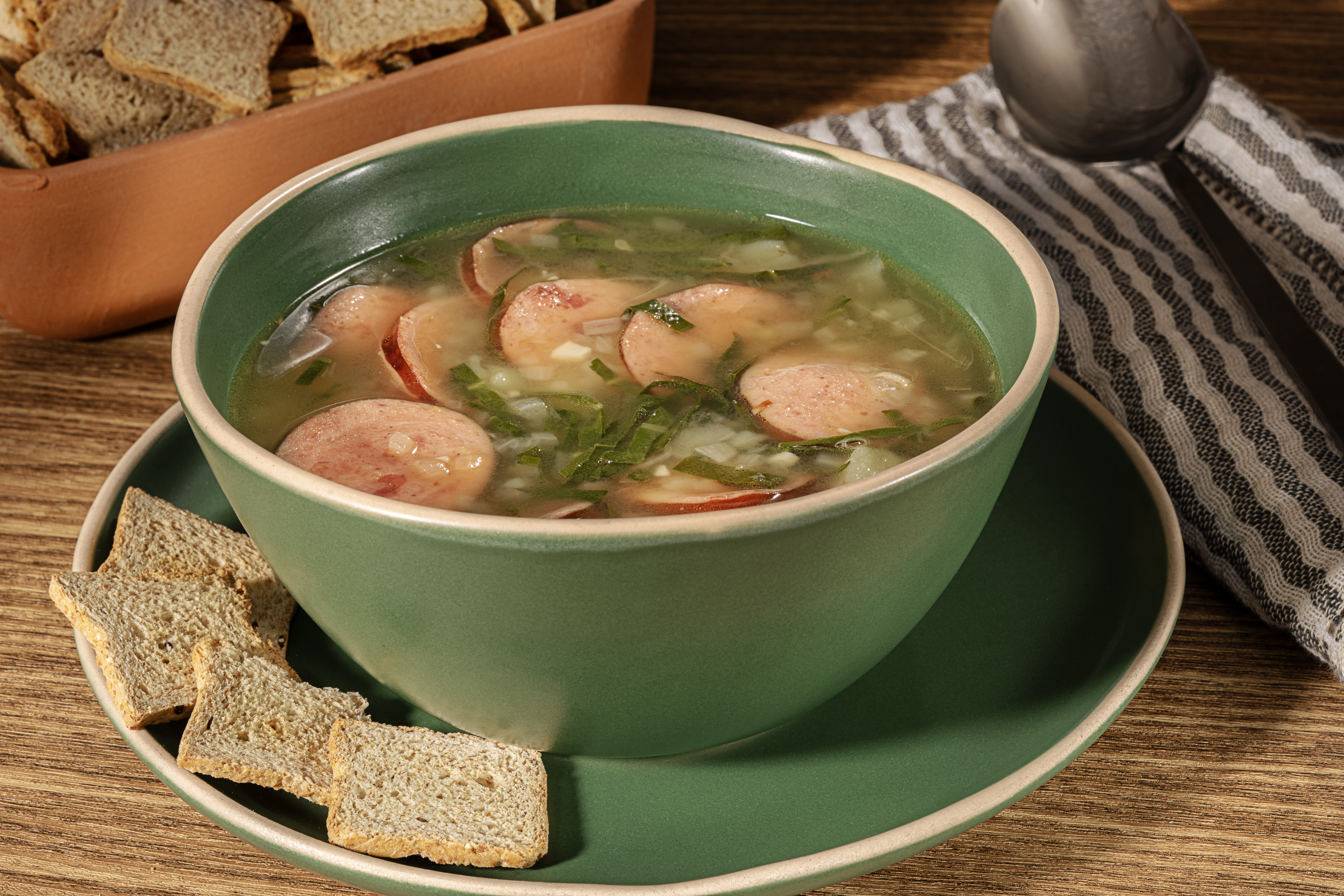 Caldo verde com rodelas de linguiça e folhas verdes em uma tigela verde, acompanhado de torradas integrais e uma colher sobre um pano de prato listrado, tudo sobre uma mesa de madeira