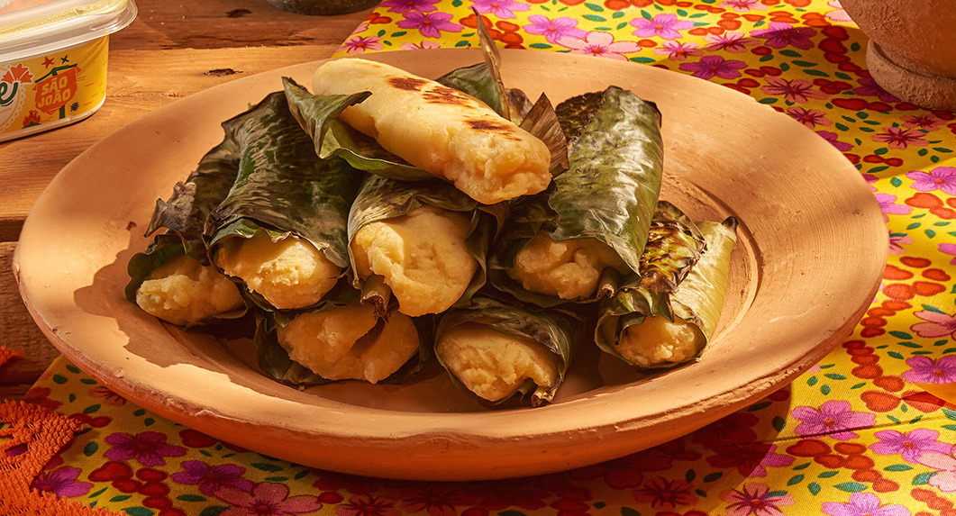 Pamonhas de milho cozidas em folhas verdes, algumas com marcas de grelha, empilhadas em um prato de barro rústico. Ao fundo, um pote de margarina e um tecido amarelo com estampas florais coloridas.