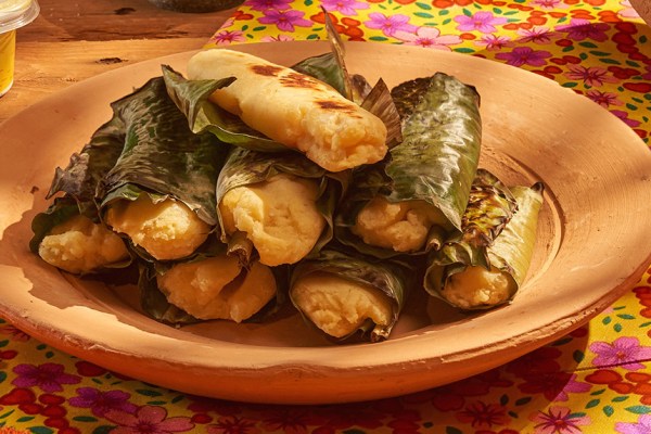Pamonhas de milho cozidas em folhas verdes, algumas com marcas de grelha, empilhadas em um prato de barro rústico. Ao fundo, um pote de margarina e um tecido amarelo com estampas florais coloridas.