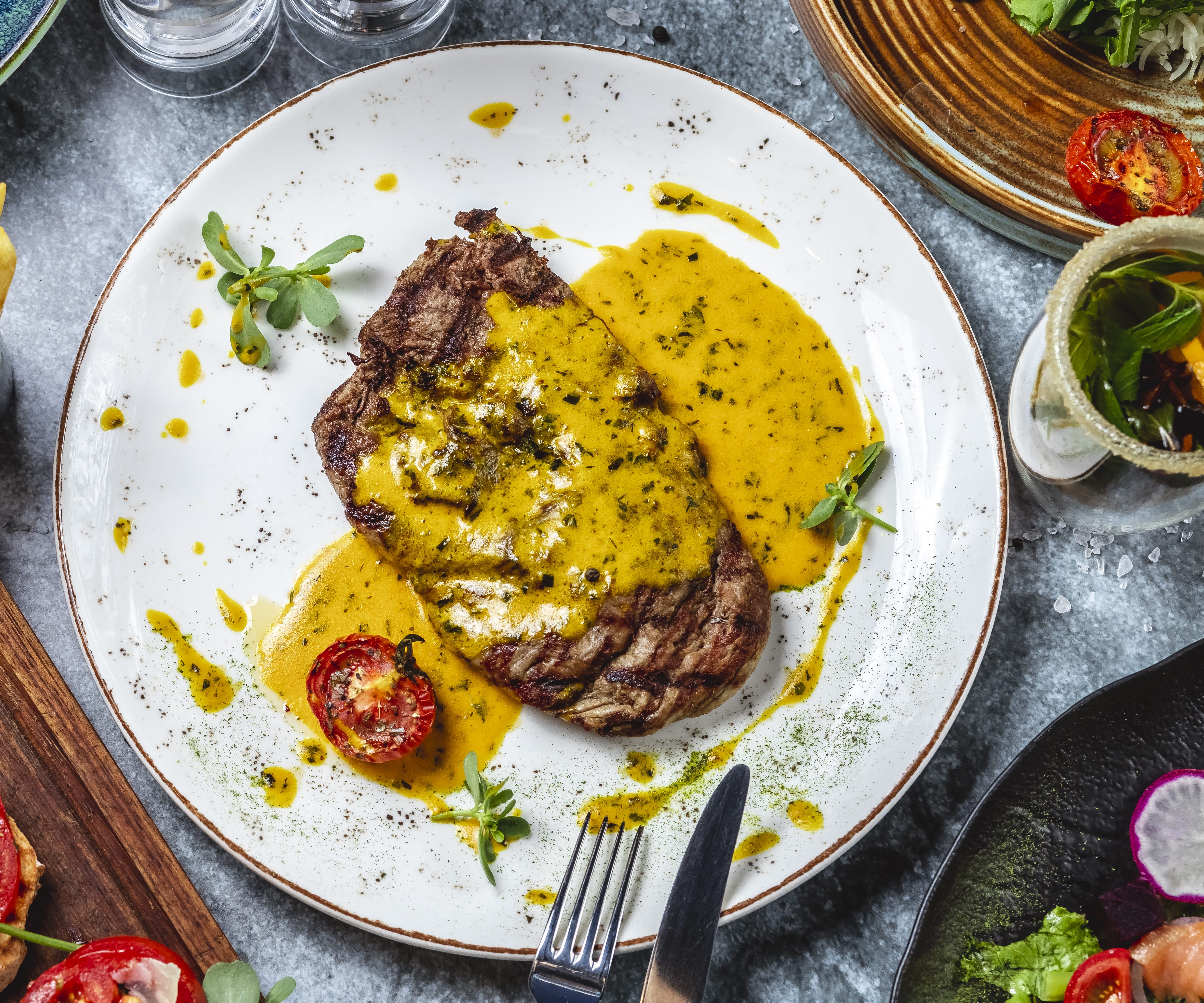 Um bife grelhado com molho amarelo cremoso e ervas, servido em prato branco. Ao lado, batatas fritas em um copo de metal e bruschettas de tomate com queijo em uma t&aacute;bua de madeira. Talheres de metal est&atilde;o pr&oacute;ximos ao prato principal